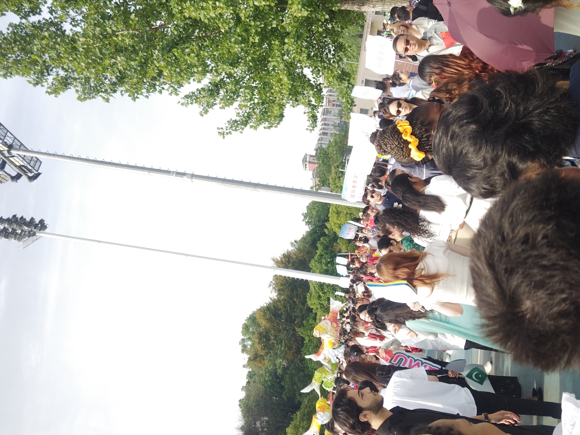 A crowd of people, mostly in white shirts, stand in a line. Some hold signs and flags, including a Pakistani flag. Overhead are trees, stadium lights, and a building in the background. The scene is in Chongqing High-tech Zone, China.