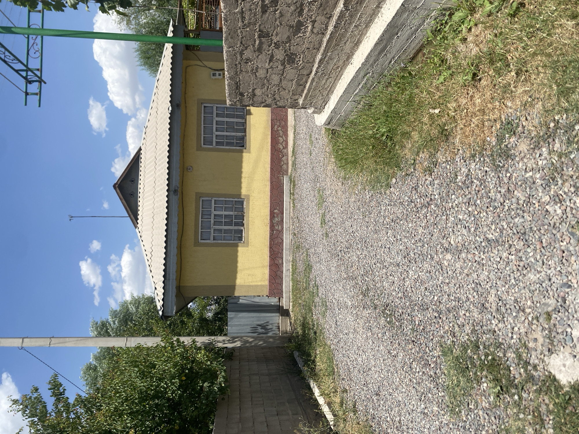 The image depicts a single-story yellow house with a corrugated metal roof, situated in a rural or suburban area. The house has two visible windows with white frames and translucent curtains. A decorative red base runs along the lower part of the house's exterior wall. The area in front of the house is a gravel driveway. To the left of the driveway is a concrete block wall, and further to the left are trees. A utility pole is visible behind the concrete wall. To the right of the driveway is a low, weathered brick structure with a concrete base, and some dry and green grass. A green metal support structure, possibly for a clothesline or awning, is visible against a bright blue sky with scattered white clouds. A thin metal fence or gate is seen between the concrete wall and the house. The overall scene suggests a sunny day. The location of Боғистон, Tajikistan is indicated, suggesting this is a property within that region. There are no people or animals visible in the image.