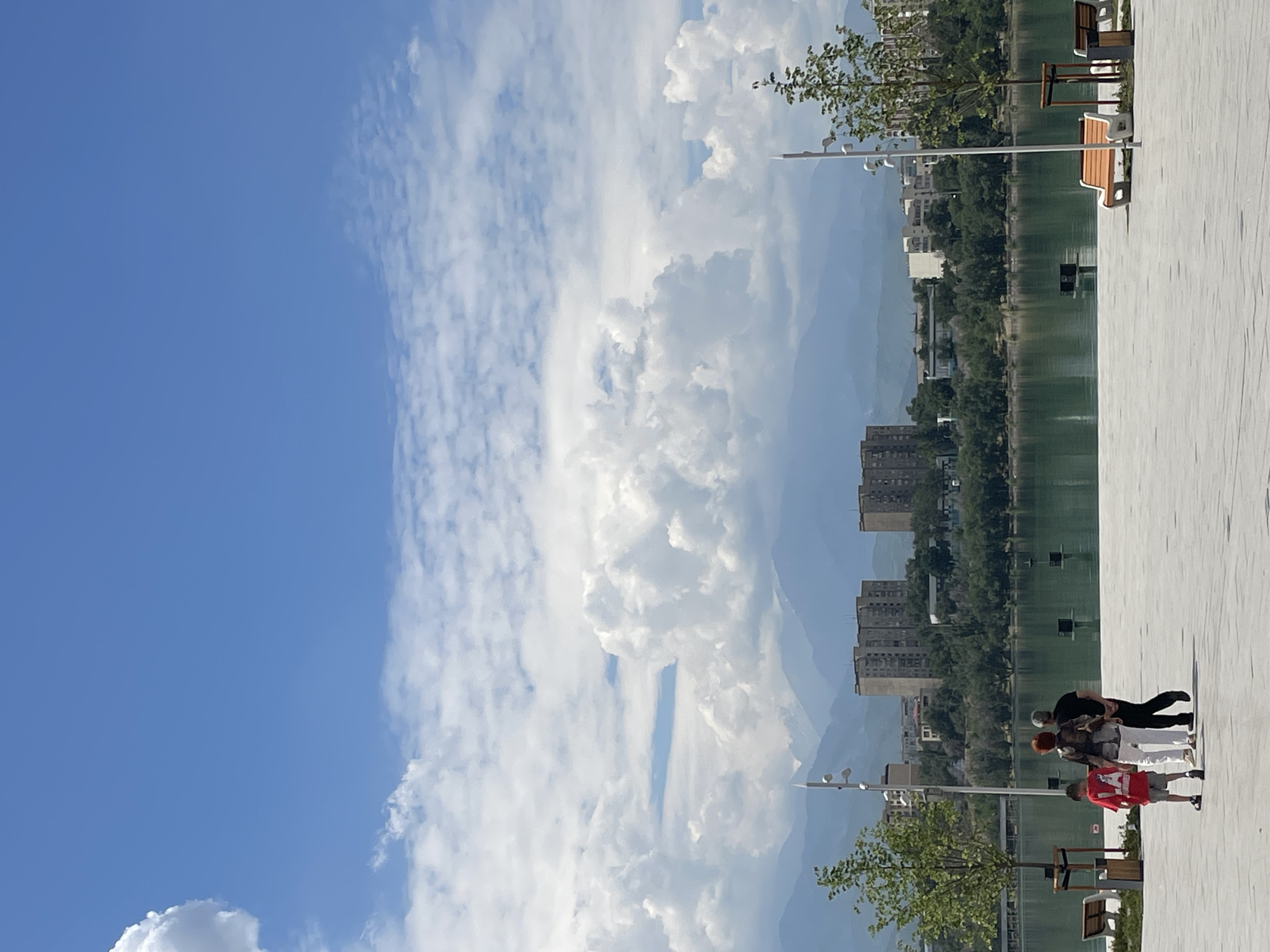 The image captures a bright, daytime scene at the Bolshoi Almaty Lake, located in Almaty, Kazakhstan. The foreground features a paved walkway with three people walking away from the camera: a young boy in a red shirt and grey shorts, an adult woman in a black and white patterned shirt and white pants, and an adult man in a black shirt and pants. To the right of the walkway, two modern park benches, one orange and white, are visible. A young tree in a planter box is situated between the benches and a lamppost. The midground shows the tranquil surface of Bolshoi Almaty Lake, reflecting the sky and surrounding landscape. The far shore is lined with lush green trees, behind which several multi-story apartment buildings are visible. In the background, a range of majestic mountains, partially shrouded in haze, rises against a vibrant blue sky filled with fluffy white clouds. The overall atmosphere is serene and picturesque, showcasing a blend of urban development and natural beauty.