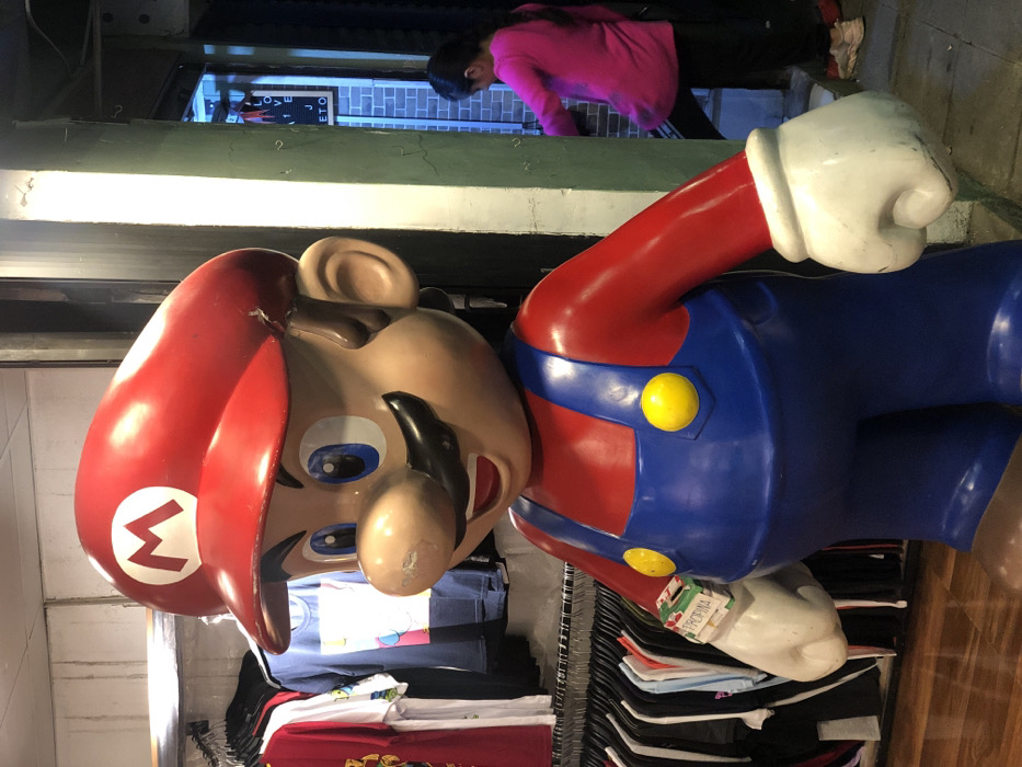 A plastic Mario statue stands beside racks of clothing. Mario wears his iconic red hat with a white "M" emblem, blue overalls, and a red shirt. A woman in a pink shirt is partially visible in the background, looking down at something. The scene is brightly lit, possibly inside a store.
