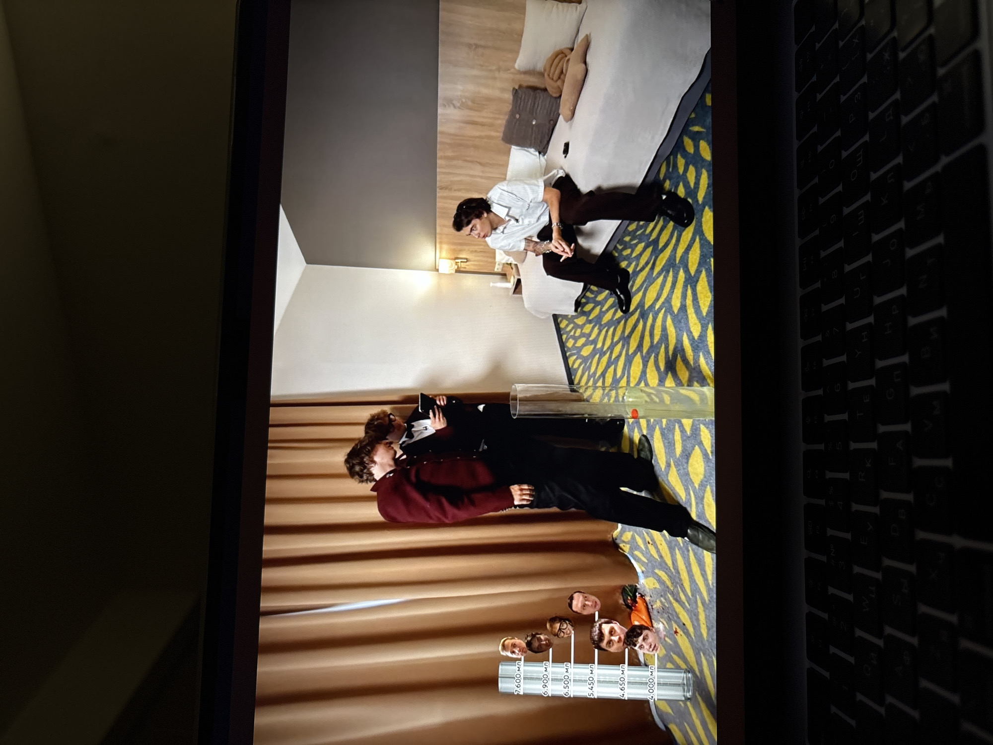 The image shows a laptop screen displaying a scene that appears to be a room, possibly a hotel room, with a patterned rug and a bed. In the foreground, on the left side of the screen, there is a tall cylindrical object with various faces attached to it at different levels, resembling a gauge or a measuring tool. Text with numbers and Cyrillic letters is visible on this object. To the right of this object, two young men in formal attire are standing. One is wearing a burgundy jacket and black pants, while the other is in a black suit and bow tie, holding a tablet or phone. On the right side of the screen, another young man is sitting on the bed, looking away from the camera. He is wearing a striped shirt and dark pants. The overall scene appears to be a staged or recorded event, as indicated by the presence of the faces on the measuring device and the composition of the shot. The text "Perm, Russia" is not present in the image itself, and therefore cannot be included in the description of the scene.