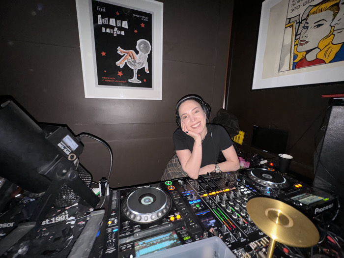 A medium-shot photograph captures a female DJ positioned behind a professional mixing console in a club or bar setting, identified as being in Moscow, Russia. The central figure is a woman with light skin and dark hair, wearing black over-ear headphones, a black short-sleeved top, and a dark plaid skirt. She is smiling directly at the camera, resting her right hand under her chin and her left forearm on the DJ equipment. A silver-toned watch is visible on her left wrist.
The DJ setup consists of two Pioneer CDJ-style players positioned on either side of a central Pioneer DJM mixer, all featuring multiple illuminated controls, faders, and rotary knobs. A small, gold-colored cymbal-like accessory is mounted on a stand to the right of the mixer. Various cables are visible connecting the equipment. To the far left, a black theatrical lighting fixture, marked "WTF-100" and "EST.1991", is angled away from the camera.
The background wall is dark brown or grey. Two framed artworks are displayed: on the left, a black poster with white Russian text "ГЛАВА КЛАБА" (Head of the Club) and "КИКИ ШОУ с ЖИВОЙ МУЗЫКОЙ" (KIKI SHOW with LIVE MUSIC), red stars, and an illustration of human legs emerging from a cocktail glass. On the right, a pop art style image depicts a blonde woman and a man in a comic book aesthetic with Russian text in speech bubbles. Behind the DJ equipment, a small monitor, a black cup, and a yellow object with visible Russian text "ГЛАВА" (GLAVA) are present.