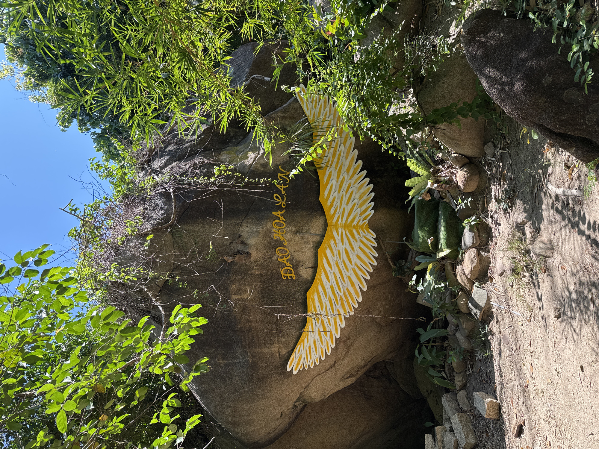 The image depicts a large rock face in Hoa Thang, Vietnam, adorned with a painted yellow and white wing motif and text in yellow lettering that reads "ĐẢO HOA LAN". The rock is situated in a natural environment with lush green foliage and trees overhead, suggesting an outdoor location, possibly a tourist attraction or a natural landmark. The ground in front of the rock is covered with dirt, pebbles, and some scattered stones. In the lower portion of the image, there are several green steps leading up to a moss-covered area in front of the rock, possibly part of a pathway or an altar-like structure. The lighting indicates a sunny day with shadows cast by the surrounding vegetation. No people are visible in the frame.