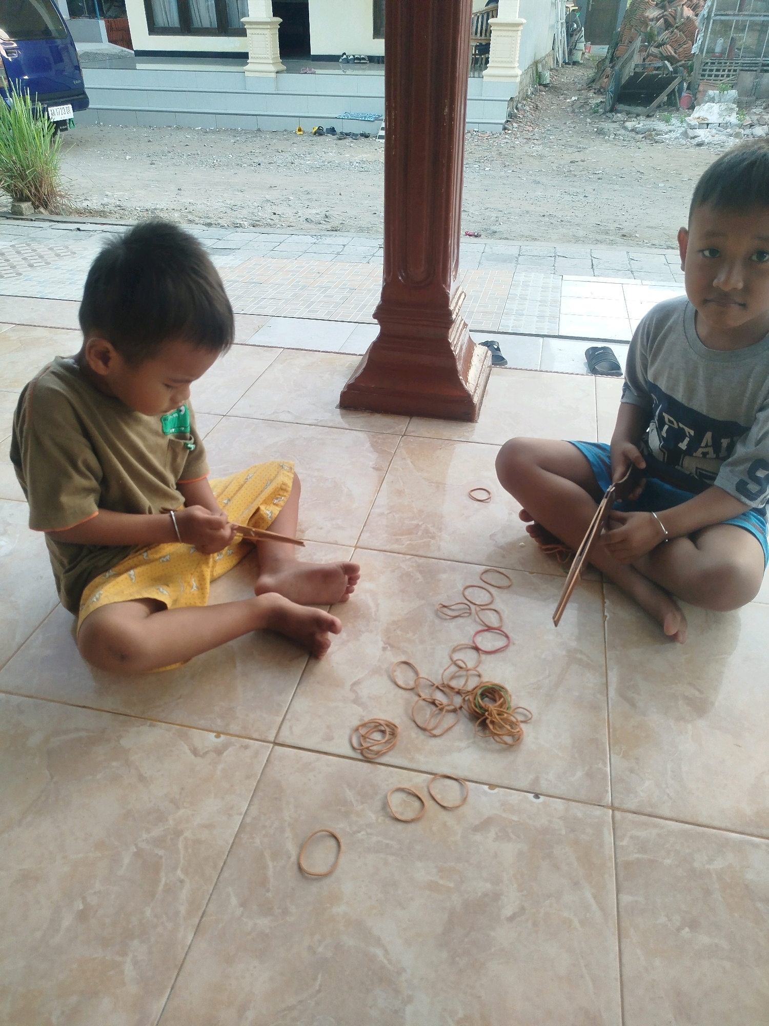 Two young boys are sitting on a tiled floor in what appears to be an outdoor covered area. The boy on the left is wearing a green t-shirt and yellow shorts with a pattern of ducks. He is holding a wooden stick and appears to be playing with a bundle of rubber bands on the floor in front of him. The boy on the right is wearing a gray t-shirt with blue accents. He is also sitting cross-legged and is holding two wooden sticks, seemingly in the process of making something with rubber bands. Scattered around the boys are numerous rubber bands of various colors. In the background, there is a blue vehicle, a portion of a building with a white pillar, and a dirt/gravel area with some debris. The setting suggests a residential area, possibly in Burneh, Indonesia, given the architectural style and general environment.