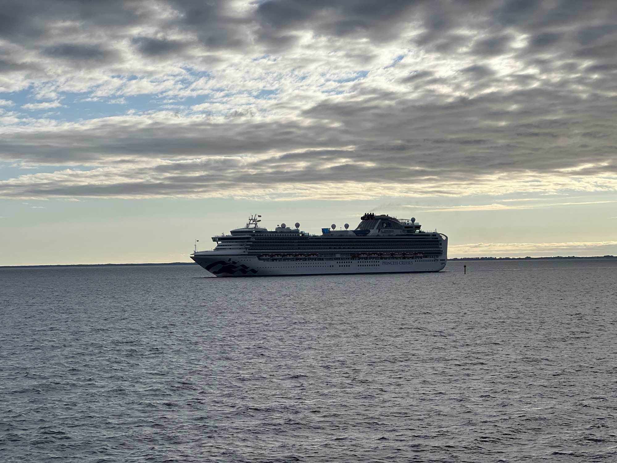 The image shows a large cruise ship, the "Emerald Princess" of Princess Cruises, sailing on a body of water under a cloudy sky. The ship is white with dark blue and gray patterns on its hull and features multiple decks with windows and railings. Smoke or steam is visible emanating from a stack on the upper rear of the ship. The water is choppy, with visible waves. A red and white buoy is present in the water to the right of the ship. The horizon line is visible in the distance, separating the water from a pale, hazy sky with layers of clouds. The scene is devoid of any specific landmarks that would indicate the city of Gdynia, Poland.