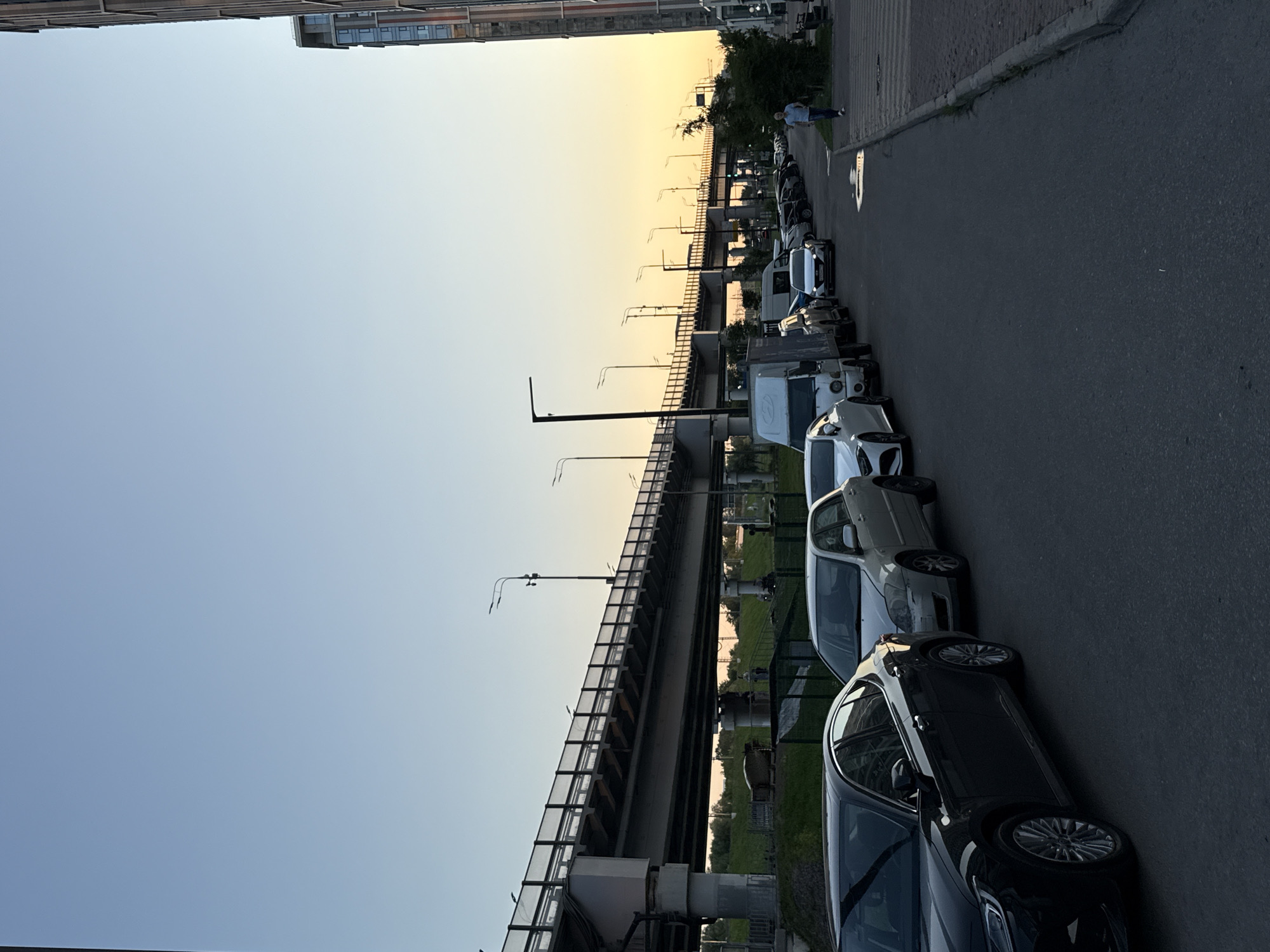 This is a vertical, eye-level, outdoor medium shot taken during the golden hour. The image captures a street scene in Khimki, Russia, with a row of parked cars lining the asphalt road. The cars are mostly sedans and a few vans, all facing away from the viewer. A pedestrian, dressed in a light blue shirt and dark pants, walks along the sidewalk.
In the background, a modern apartment building stands tall on the left, contrasting with the sky that transitions from a soft blue to a warm yellow at the horizon, indicating sunset. A multi-level overpass stretches across the scene, its concrete structure and railings visible. Streetlights are spaced along the overpass, and some appear to be on. Green grass and trees are visible on the right side of the overpass. The overall mood is peaceful and serene, bathed in the warm glow of the setting sun.