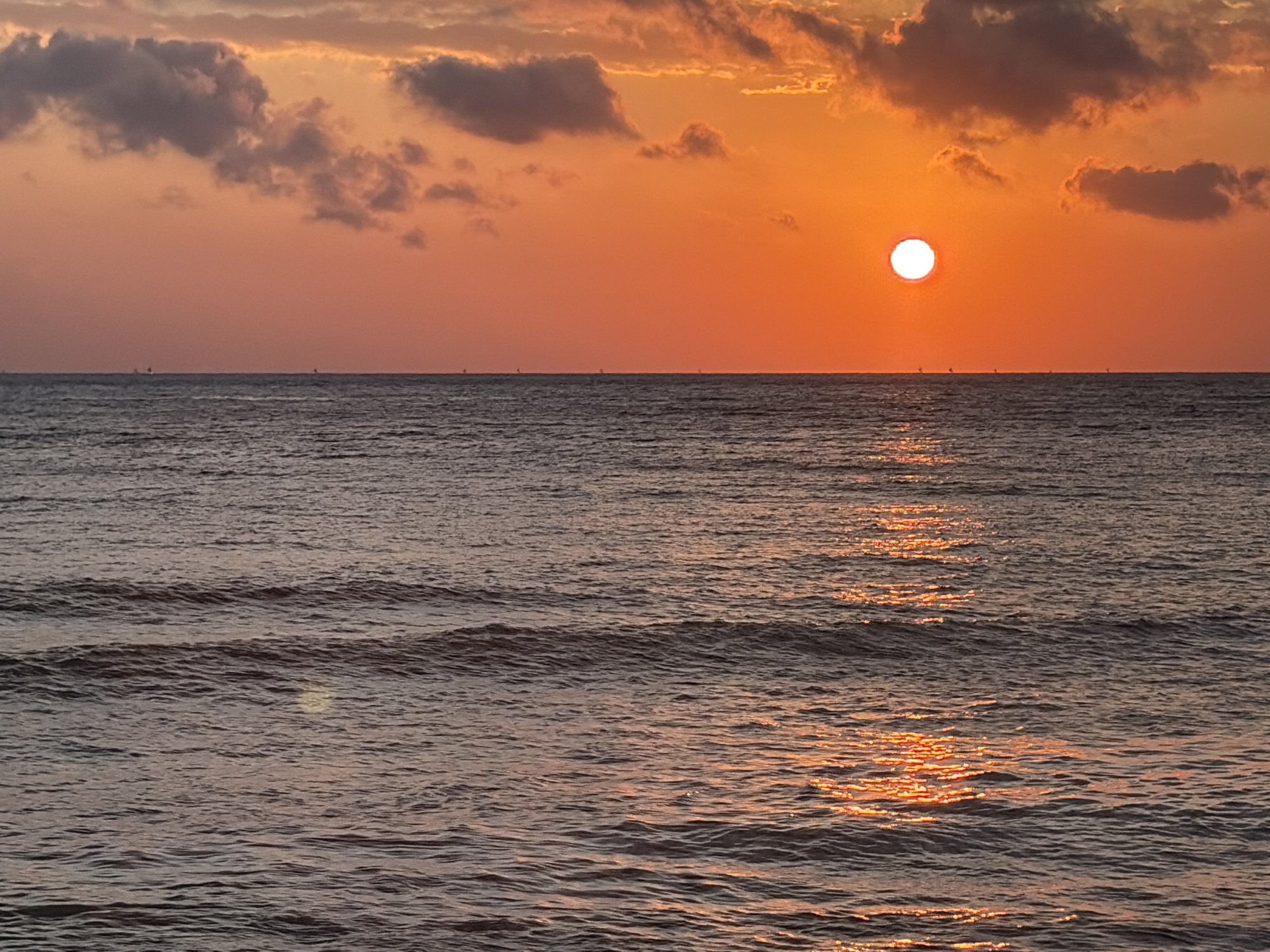 A low-angle, wide shot captures a serene ocean sunset over the waters near Abang, Indonesia. The sky is painted in vibrant hues of orange and pink, with scattered dark clouds tinged with orange light. The sun, a bright white disc, is positioned slightly left of center in the upper third of the frame. The calm ocean surface reflects the fiery colors of the sky, creating shimmering paths of light across the dark, rippling water. In the distance, along the horizon, several small, dark silhouettes of sailboats are visible. The scene is devoid of any people or specific landmarks that would identify a precise location within Abang.