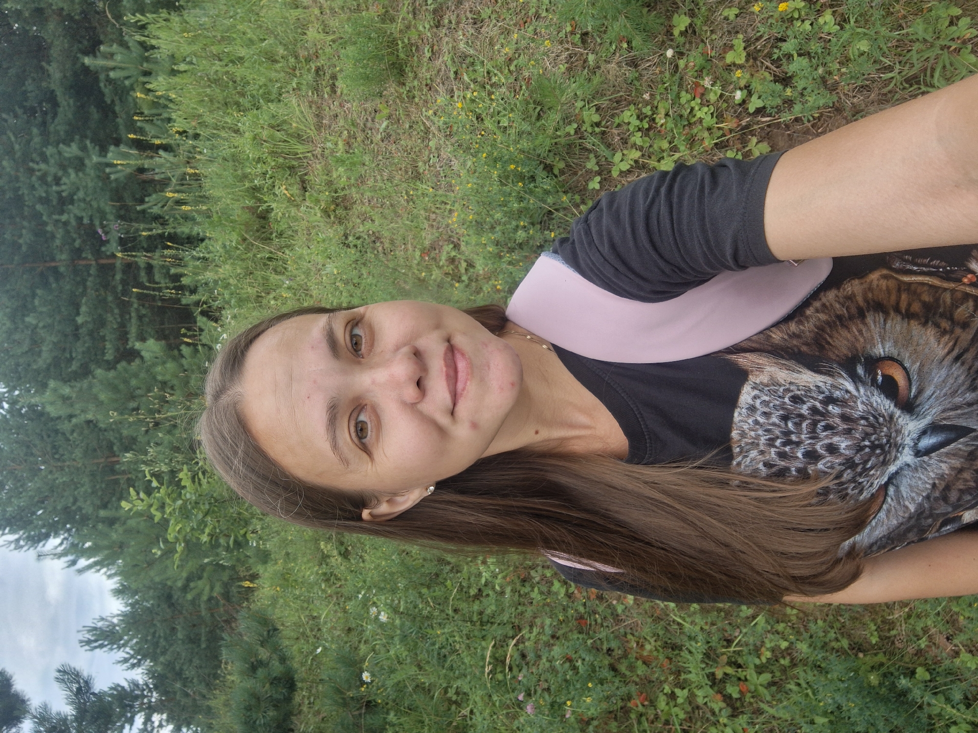 A young woman with long brown hair and a light complexion is taking a selfie outdoors. She is wearing a black t-shirt with a large graphic of an owl's face and a pink backpack strap visible over her shoulder. She is smiling faintly. The background consists of a grassy slope with scattered wildflowers and trees, suggesting a rural or natural setting. There are no visible indications of urban environment or specific landmarks of Malyi Bukor, Russia.