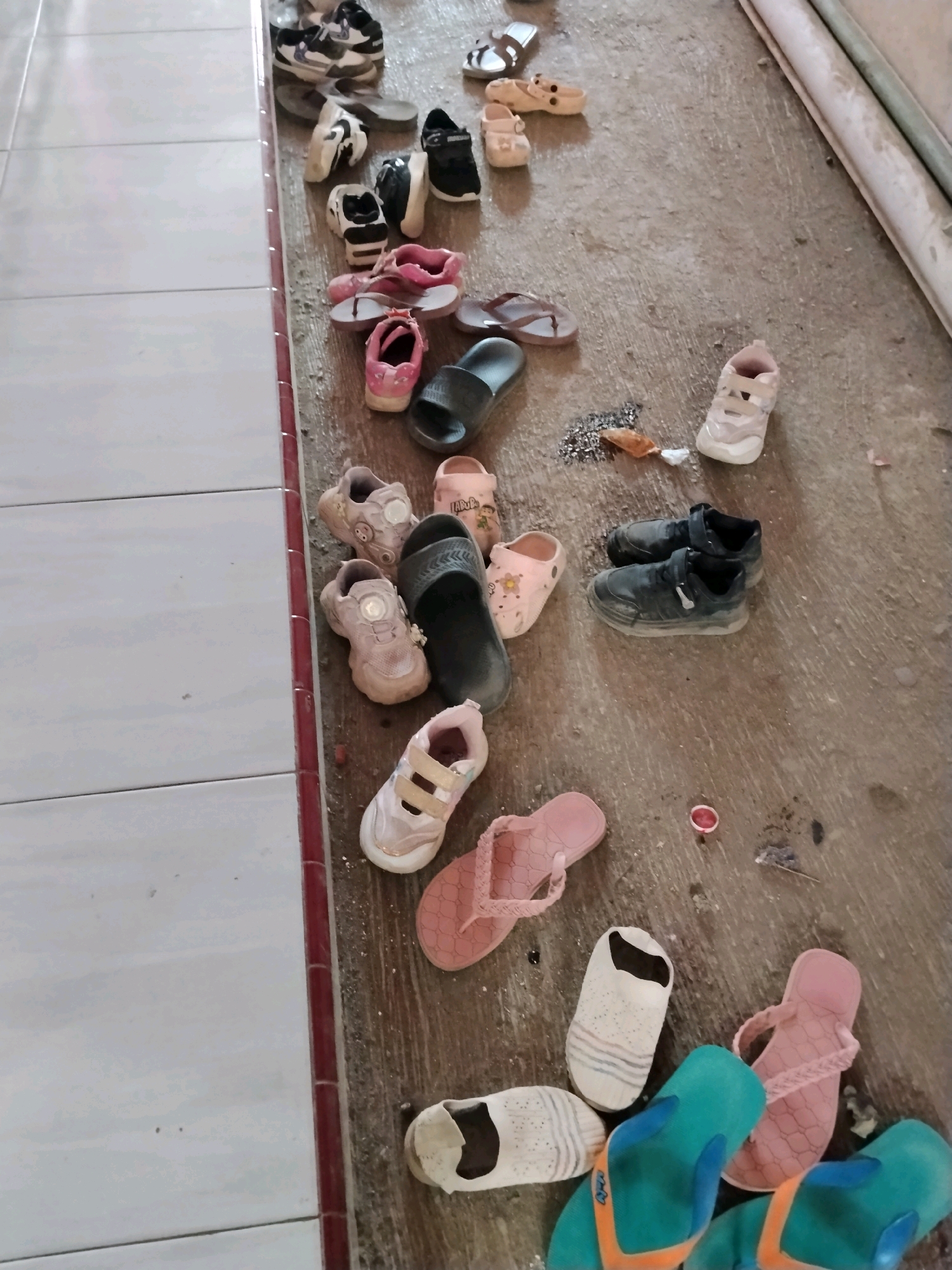 In Panggungan, Indonesia, a collection of children's footwear is scattered on a dirty concrete floor adjacent to a tiled surface. The shoes include various pairs of sandals, sneakers, and clogs in colors such as pink, black, white, and blue. Some items appear worn. The environment suggests a casual or outdoor setting, possibly a shoe rack area or entryway.