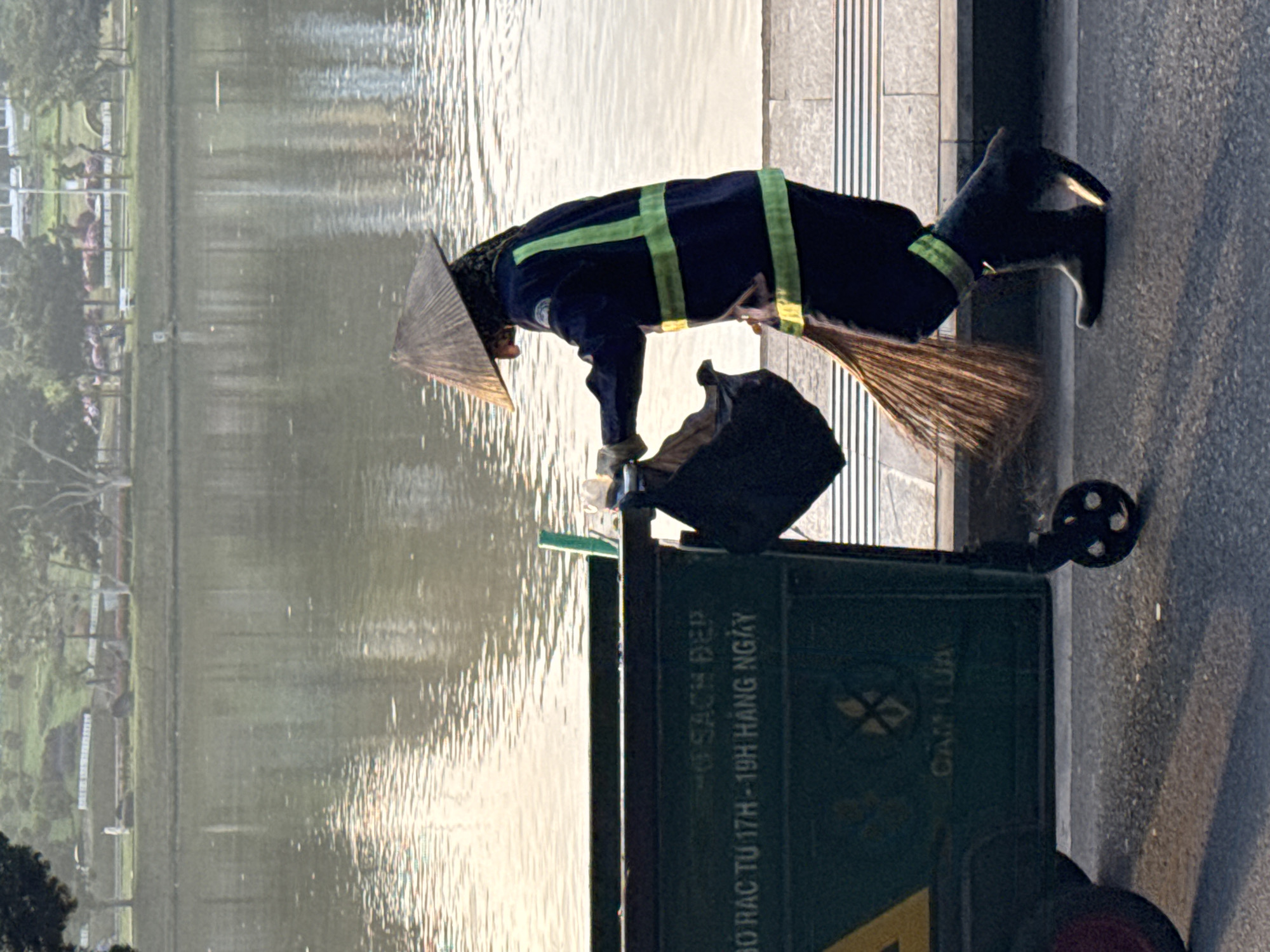 A street cleaner wearing a conical hat and a dark blue uniform with neon green reflective stripes is pushing a green refuse bin along a paved walkway next to a body of water. The cleaner is holding a broom and appears to be sweeping. The refuse bin has text in Vietnamese that reads "BO RAC TU 17H - 19H HANG NGAY" which translates to "Garbage collection from 5 PM - 7 PM daily" and "CAM LUA" meaning "No fire". The background shows a calm body of water reflecting trees and buildings, suggesting a park or lakeside area. The scene is likely in Da Lat, Vietnam, given the conical hat and Vietnamese text.