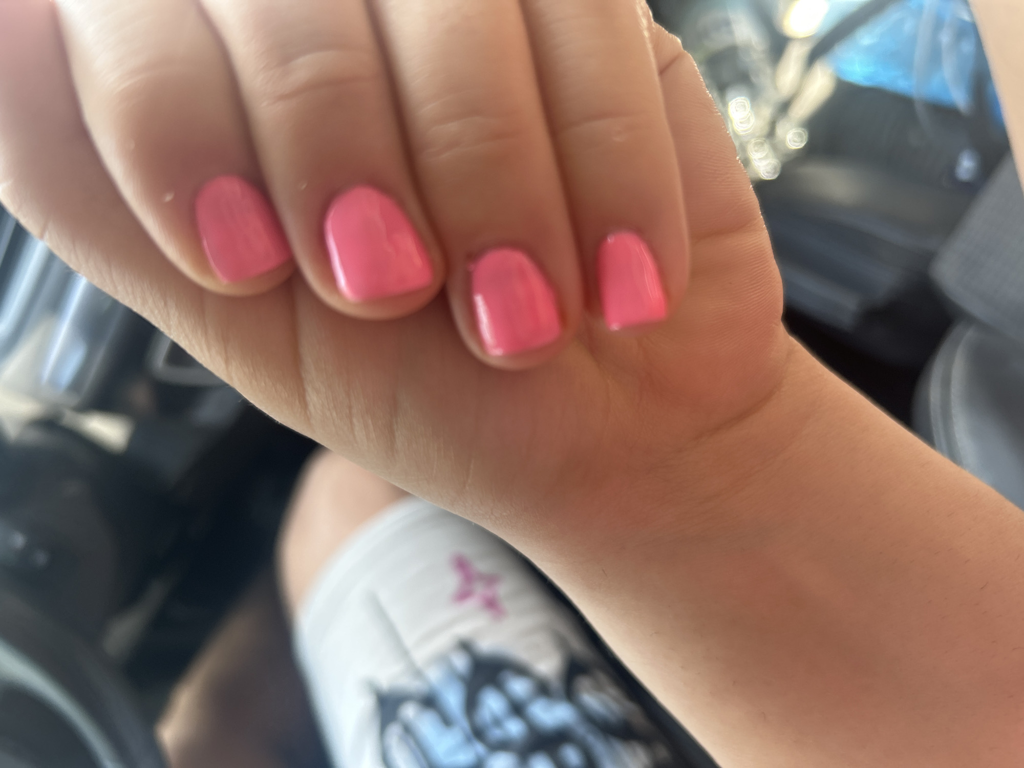 The image shows a close-up of a person's hand with bright pink painted nails. The hand is in the foreground, and out-of-focus elements of what appears to be a car interior are visible in the background, including a steering wheel and a dashboard. A portion of white fabric with a black and blue pattern and a small pink detail is visible on the left side of the image, which could be clothing or an accessory. The lighting suggests it is daytime. There is no information in the image that indicates the location is Plovdiv, Bulgaria.