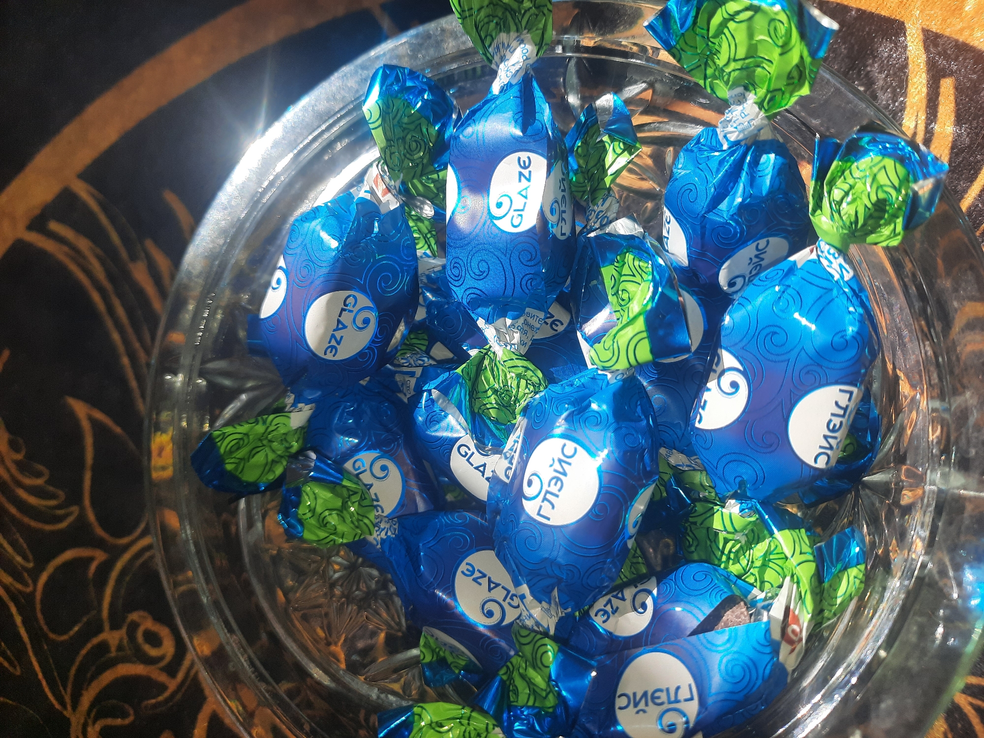 A close-up, overhead shot captures a glass bowl filled with individually wrapped candies. The candies are a mix of blue and green wrappers, many of which bear the brand name "GLAZE" or its Cyrillic equivalent "ГЛЭЙС" in white lettering. The wrappers feature a swirled pattern. The glass bowl rests on a surface with an ornate, dark background, possibly a tablecloth with a metallic sheen. The lighting creates highlights and shadows, giving the scene a slightly dramatic effect. There are no people present in the image. The location in Shklov, Belarus is not evident from the image itself; this detail is external information.