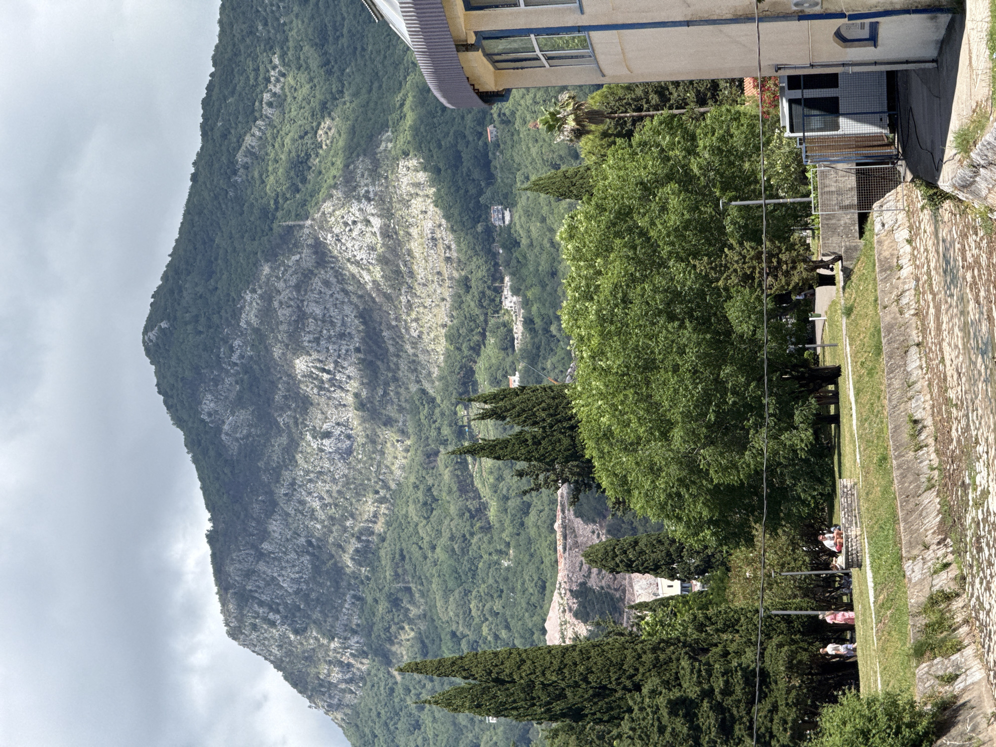 This daytime scene captures a panoramic view of a mountainous landscape, likely near a town or city in Budva, Montenegro. The left side of the frame is dominated by a majestic, steep mountain, densely covered in green trees, with lighter grey rock faces exposed in places. The sky above is overcast but bright.
In the mid-ground and right foreground, there is a built-up area and a green, park-like space. A prominent, light-colored building with a corrugated roof and blue accents, including window frames and a stripe, is situated in the upper right. A dark blue symbol, possibly an 'H' or stylized 'N' within a square, is visible on its facade.
Below this building, a paved area leads to a fenced enclosure. Along the lower right, a textured stone wall or embankment runs through the scene, bordered by a strip of green grass. Tall, slender cypress trees are scattered throughout the mid-ground, alongside other broad-leaved trees and a palm tree, indicating a Mediterranean climate.
A few people are present in the lower right section of the image. One person is standing in light clothing, while a small group is seated at a stone table with benches under the shade of trees. Another individual is visible further left among the foliage. Beyond the immediate foreground, the mountainous terrain extends, revealing more distant green slopes and small, scattered buildings nestled within the landscape. The number "10" is visible on a white sign attached to a metal fence in the right foreground.