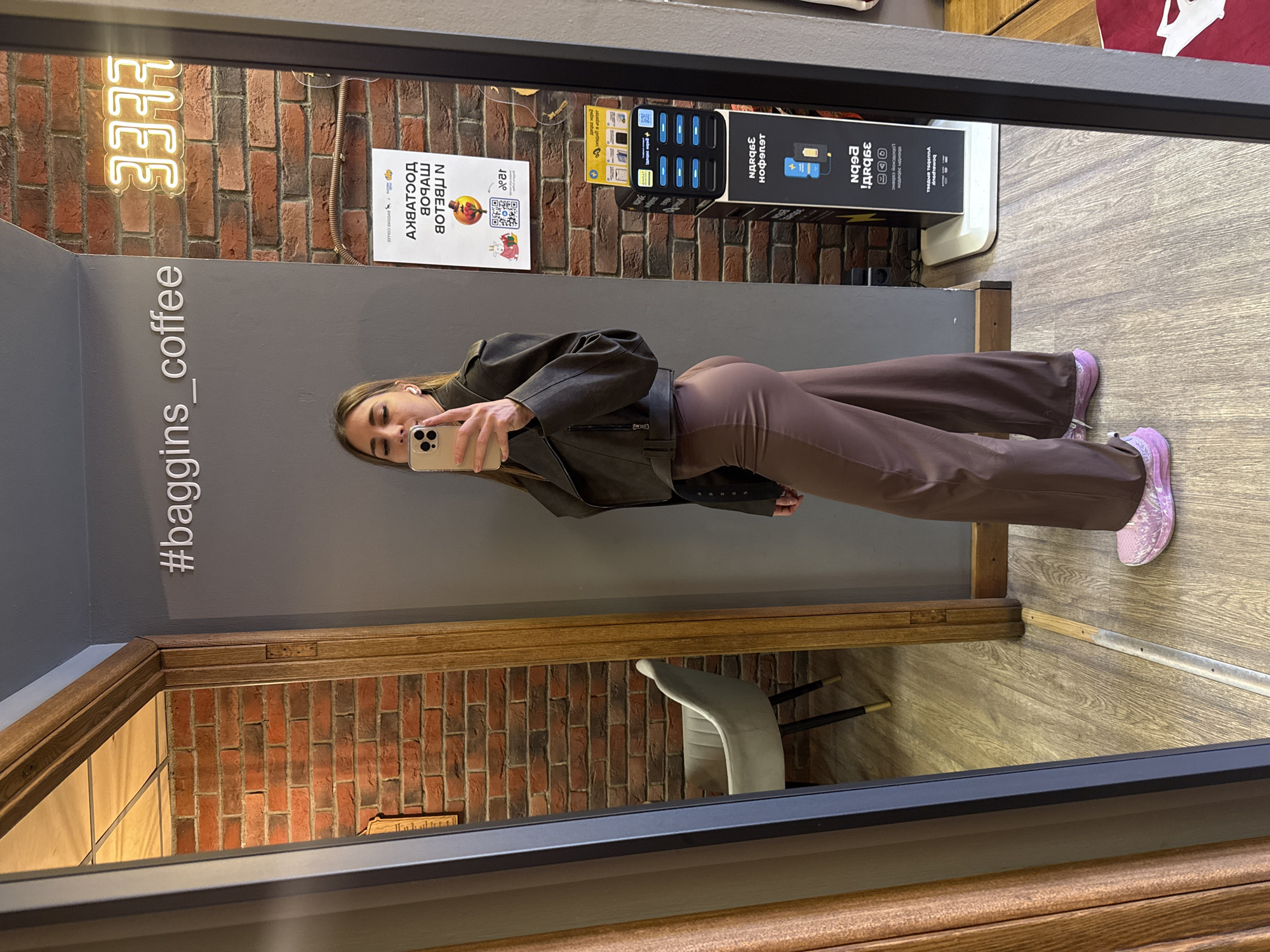 A woman is taking a mirror selfie. The mirror reflects a coffee shop interior, as evidenced by the hashtag "#baggins_coffee" in the reflection. The Russian text and a sign referencing "IZ COLLEE" further suggest this is a business in Russia. Given the hashtag, the location is most likely Saint Petersburg, Russia, where a coffee shop with that name is present.
The mirror shows a brick wall with neon signage. Other objects visible include a charging station for mobile devices and a sign with Russian text and a QR code. The woman is wearing a brown jacket, brown pants, and pink sneakers. In the reflection of the lower portion of the mirror, a chair and more of the brick wall are visible. The photograph is taken indoors, likely inside the coffee shop.