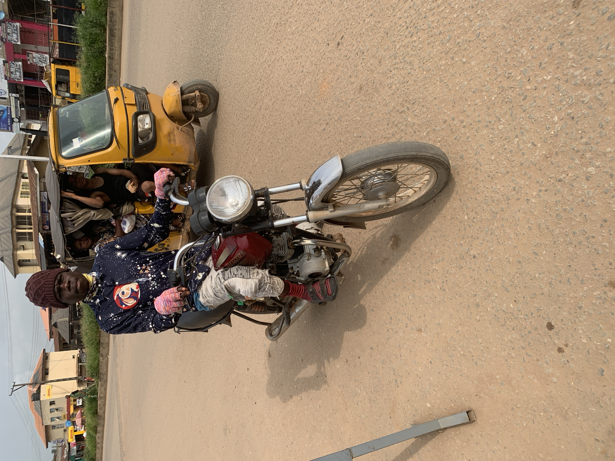 In Shagamu, Nigeria, a young man wearing a brown knitted cap and a dark blue shirt with white star-like patterns and a circular graphic sits on a dark red motorcycle. He has on pink gloves and light-colored pants with red and black striped socks and dark sandals. A yellow motorized tricycle with a dark fabric canopy is visible behind him, carrying passengers. The tricycle has a black tank top-clad male driver. The scene takes place on a paved road in front of buildings with signs, including one that reads "DHL." The environment suggests a commercial or residential area of the city.