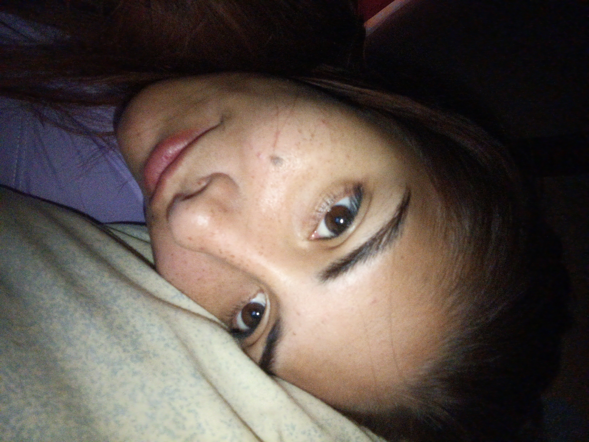 A close-up, angled portrait of a woman lying on her side. Her head is turned to face the camera. She has brown hair, light-colored skin with freckles, and brown eyes. A light-colored blanket with a subtle pattern is near her face. A lavender or light purple pillow is visible behind her head. The background appears dark and indistinct. There are no other details available to suggest the location is Juan Griego, Venezuela.