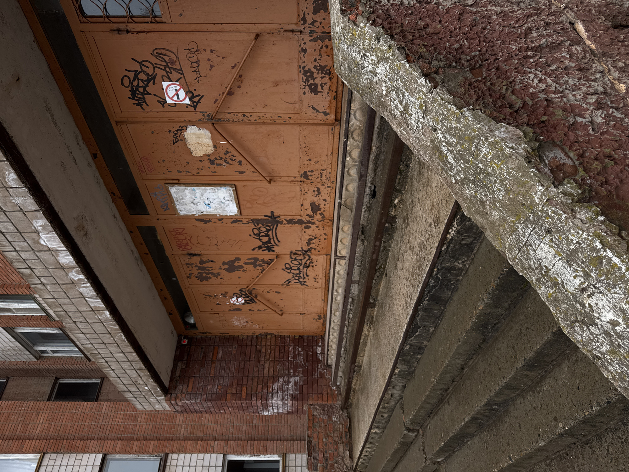 The image shows the underside of an architectural structure in Votkinsk, Russia. A rusted, orange-colored metal garage door with peeling paint and graffiti is visible. A "no smoking" sign is affixed to the door. The garage door is attached to a brick building facade. Adjacent to the garage door is a concrete structure with a heavily textured surface. The image provides no information about people, their actions, or their interactions.