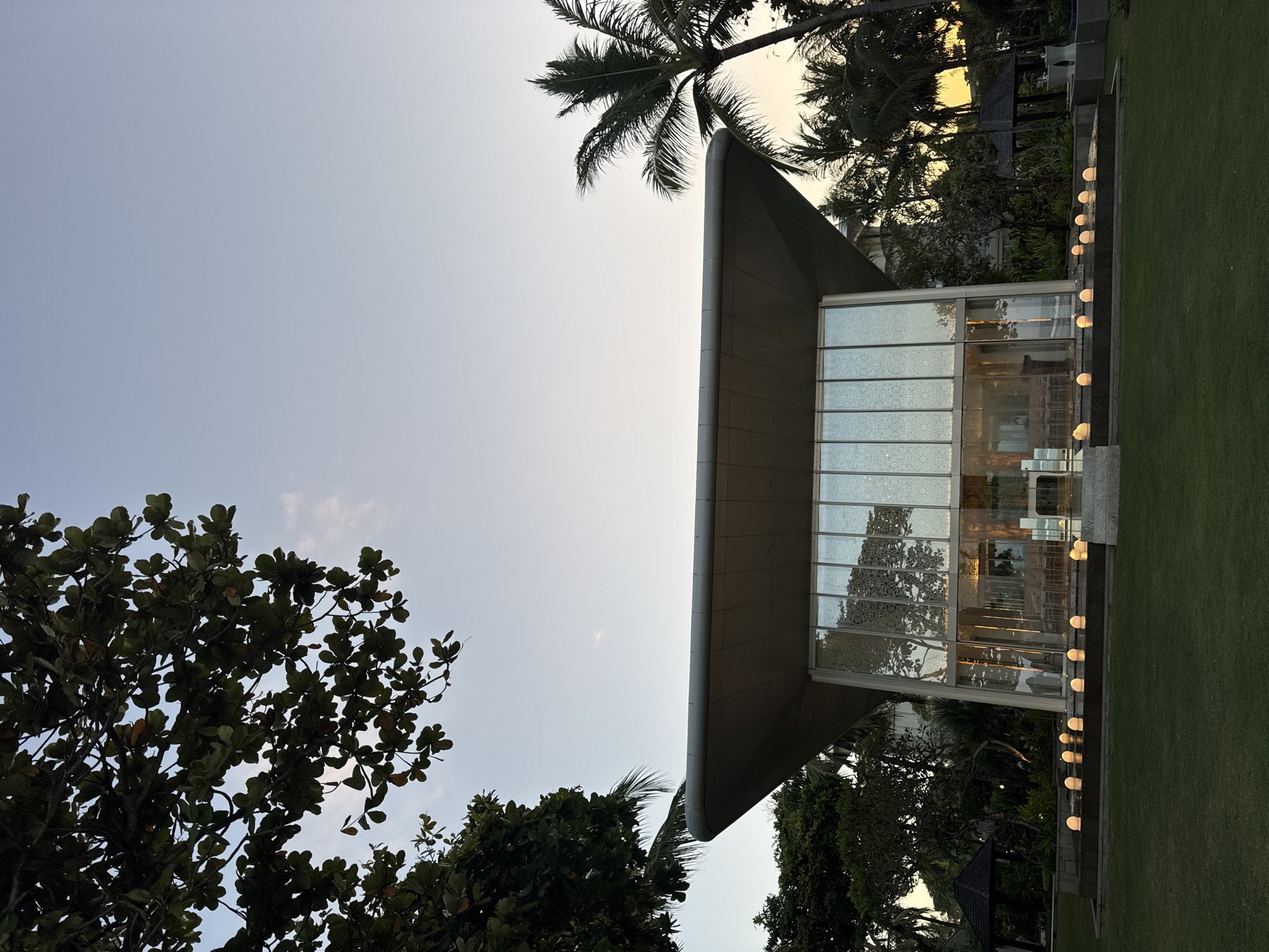 In the city of Kuta Selatan, Indonesia, a modern glass building, likely a chapel or event venue, stands at dusk. The structure features a wide, flat roof that extends outwards, supported by thin pillars, and is clad in glass panels with a patterned overlay. Inside, tables and chairs are visible, suggesting an interior space for gatherings. In front of the building, a rectangular water feature is lined with glowing, spherical lights. A lush green lawn surrounds the building, and mature trees, including palm trees, frame the scene. The sky above is a soft gradient of blue and orange, indicative of sunset. There are no people visible in the image.