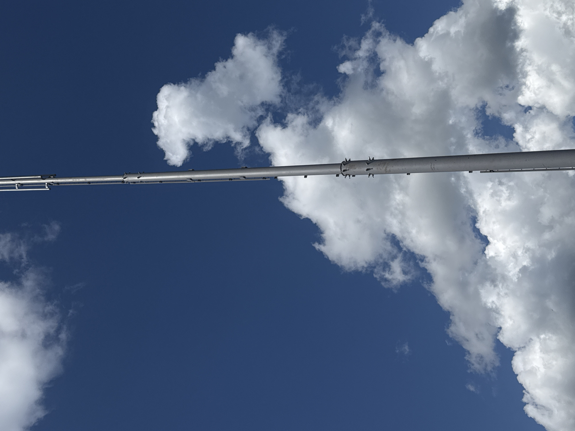 A tall, silver antenna tower stands against a bright blue sky dotted with white, fluffy clouds. The tower is segmented and appears to be constructed of metal, with visible bolts and joints along its length. The bottom of the tower is obscured by a large cloud formation, while the top extends upwards, displaying a series of vertical antennas. The image is taken from a low angle, looking up at the tower and sky. There are no people or animals visible in the scene. The location is identified as Schokino, Russia.