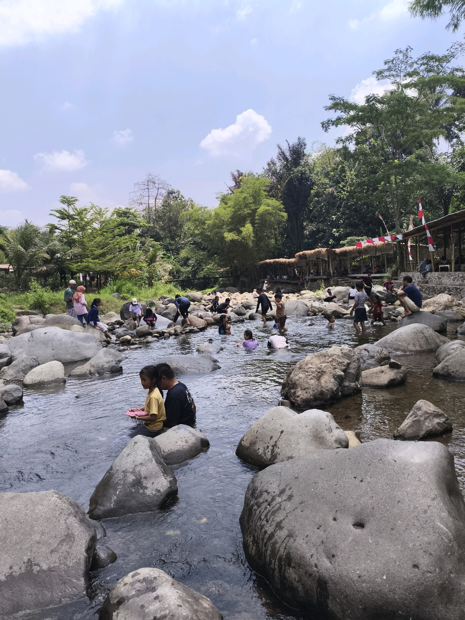 This image captures a lively scene at a river in Babakan Madang, Indonesia, where a group of people, predominantly children, are enjoying a day outdoors. The riverbed is filled with large, smooth grey rocks, some submerged in the clear, flowing water, while others rise above the surface. A diverse mix of individuals are present, with many children actively playing in the shallow parts of the river, splashing and interacting. Some adults are also visible, either supervising the children, resting on the rocks, or participating in the water activities.
The background reveals a lush, green environment with dense trees and vegetation lining the riverbanks. Several simple structures with thatched roofs are set up along the river, suggesting a recreational area or a place for vendors. Red and white flags are displayed on some of these structures, indicating a festive atmosphere. The sky above is a bright blue with scattered white clouds, suggesting fair weather. The overall impression is one of a vibrant community gathering in a natural setting.