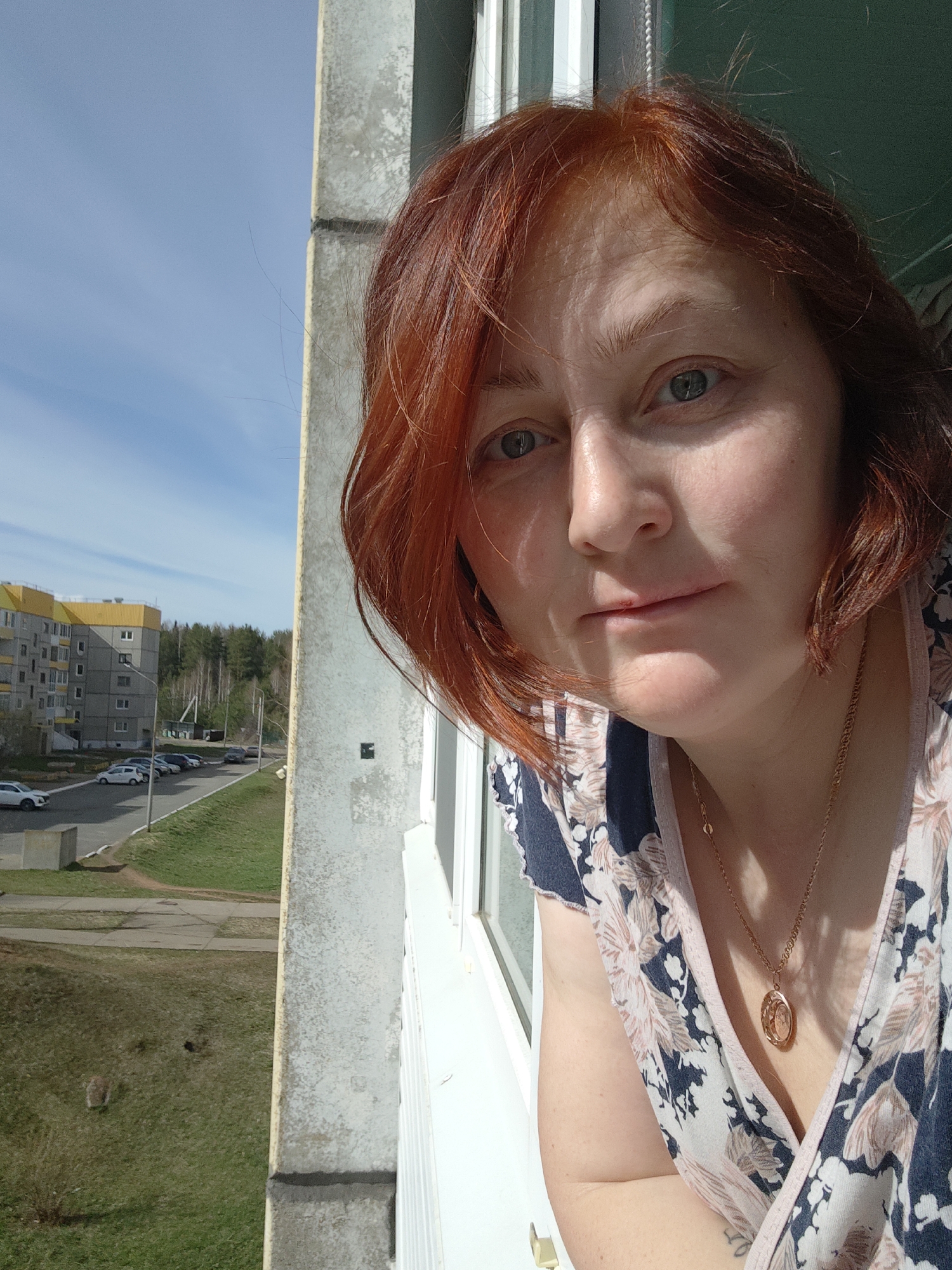 A red-haired woman stands at an open window in an apartment building in Markovskii, Russia. She is wearing a floral print shirt and a gold necklace with a pendant. Outside the window, a multi-story building with a yellow roof is visible, along with cars parked along a street, a green lawn, and trees. The sky is blue with some clouds. The window frame is white, and the building exterior is concrete.