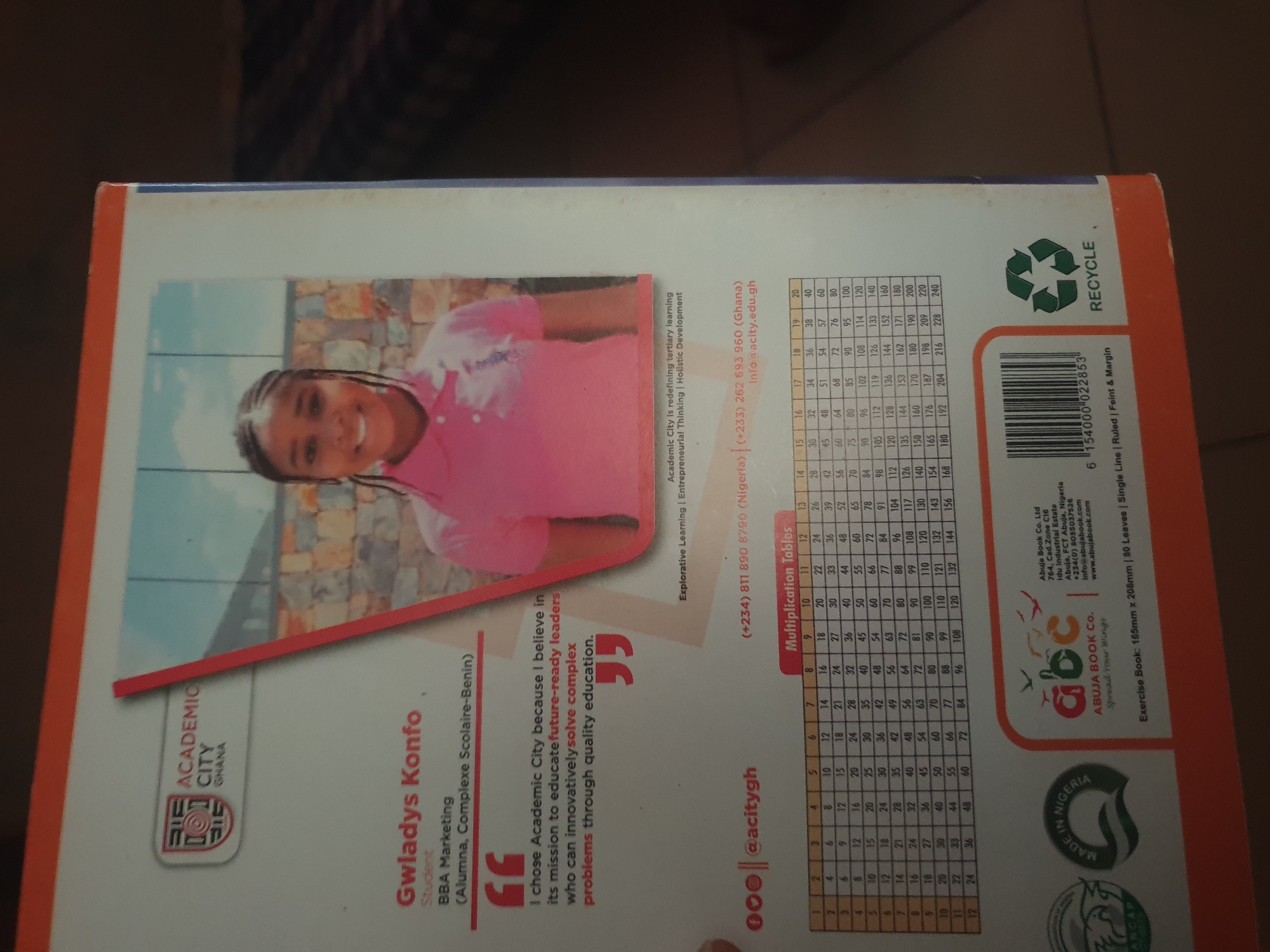This media file is a close-up shot of the back cover of an exercise book, likely a school supply. The cover features a young Black girl with braided hair, smiling warmly at the camera. She is wearing a pink collared shirt. Behind her, a blurred background suggests an outdoor setting with a stone wall and blue sky.
The cover prominently displays branding for "Academic City," an institution that appears to be located in Ghana and Nigeria. Text indicates it is a tertiary learning institution focused on "Explorative Learning | Entrepreneurial Thinking | Holistic Development." Contact information, including phone numbers for Nigeria and Ghana, an email address, and a website, is visible.
A significant portion of the cover is dedicated to multiplication tables, ranging from 1x2 up to 20x20, printed in a clear, tabular format. This suggests the exercise book is designed for students learning mathematics.
Further details include an "Abuja Book Co. Ltd" logo and contact information, identifying them as the publisher. The "Made in Nigeria" label with an emblem and the "Recycle" symbol are also visible. The product details state it's an "Exercise Book: 165mm x 208mm | 80 Leaves | Single Line | Ruled | Feint & Margin." The overall impression is that of a product for educational use, emphasizing both academic achievement and a positive brand identity.