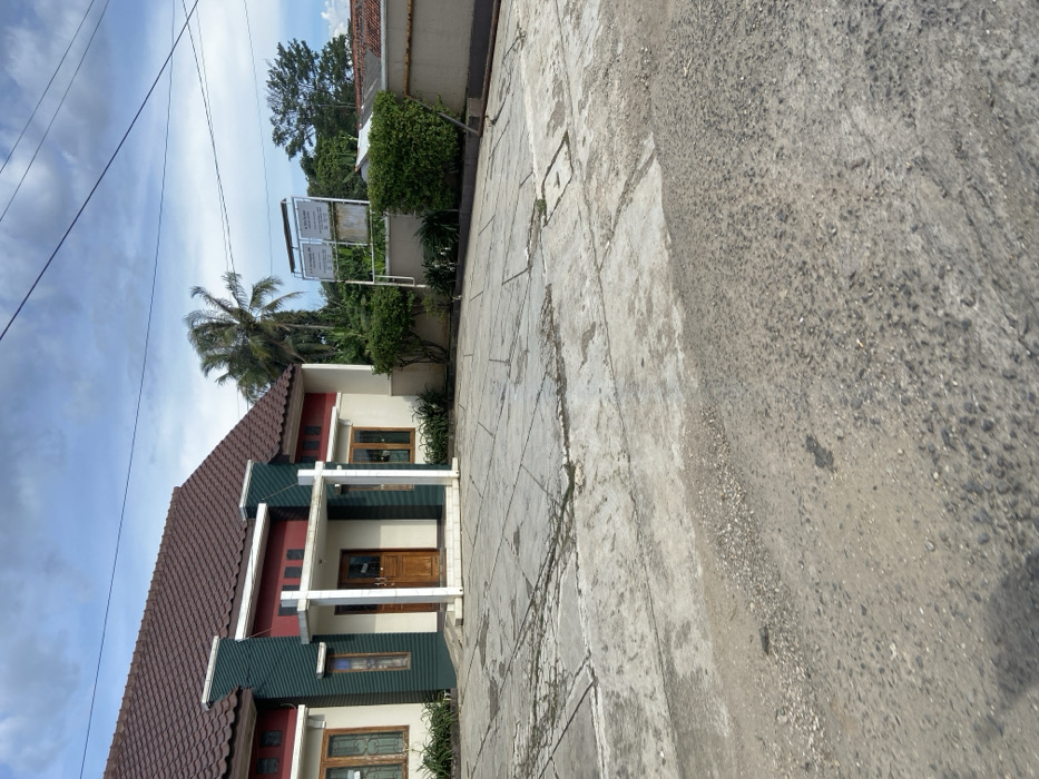 The image shows a cracked concrete driveway in front of a single-story house with a red tiled roof. The house has a green painted trim and a porch with a wooden door. The driveway is located in Sukatani, Indonesia, at coordinates -6.608 latitude and 107.414 longitude. The house is surrounded by a concrete wall with a small patch of green grass. There are several trees in the background, including a tall palm tree and some smaller trees. The sky is blue with some white clouds. The sun is shining. A metal sign is attached to the wall behind the house. The ground in the distance is gravel. There is a small metal drain in the driveway.