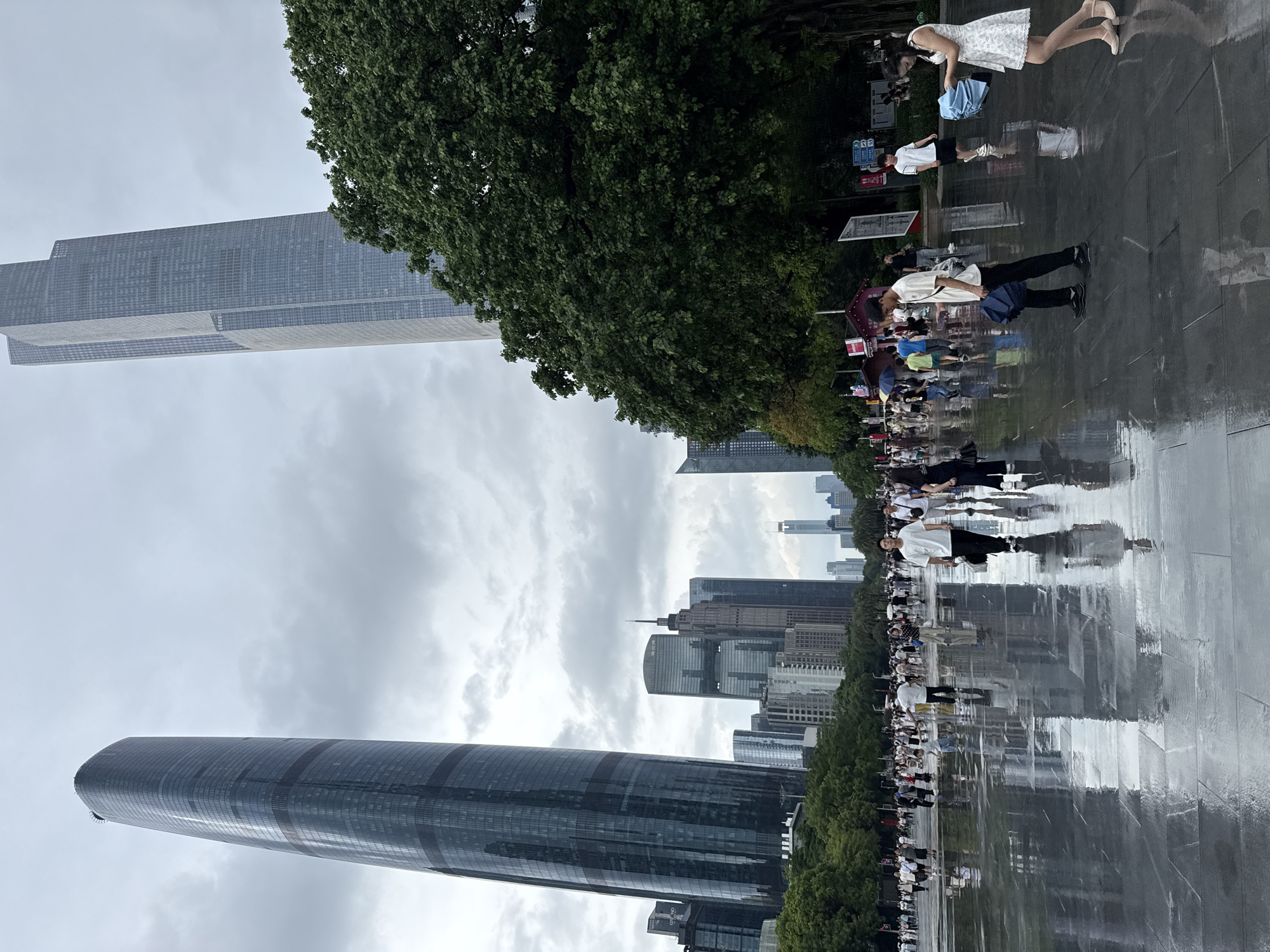 The image captures a scene in Tianhe District, China, featuring a wide, wet plaza reflecting the overcast sky and surrounding modern architecture. Dominating the skyline are two imposing skyscrapers; one on the left is a tall, curved, dark-glass building with distinct horizontal bands, while the one on the right is a more angular, lighter-toned skyscraper with a stepped profile. In the middle ground, a dense cluster of various other skyscrapers creates an urban backdrop.
The plaza's surface is wet and reflective, indicating recent rain or a cleaning. Numerous people are walking across the plaza, many holding umbrellas, suggesting continued light precipitation or damp conditions. The individuals are casually dressed, moving in various directions. Some are walking alone, while others are in small groups. A man in a white shirt and black pants walks towards the viewer, holding a dark umbrella. To his right, a woman in a white dress holds a blue umbrella. Reflections of the people and buildings are visible on the wet ground.
On the right side of the image, a large, lush green tree provides a stark contrast to the urban landscape. The area appears to be a public space, possibly a park or a large square designed for pedestrian use, with pathways and green spaces interspersed with the monumental buildings. Street signs and some smaller structures are visible along the periphery. The overall atmosphere is one of a bustling city on a cloudy day, with the wet ground adding a dynamic visual element to the scene.
