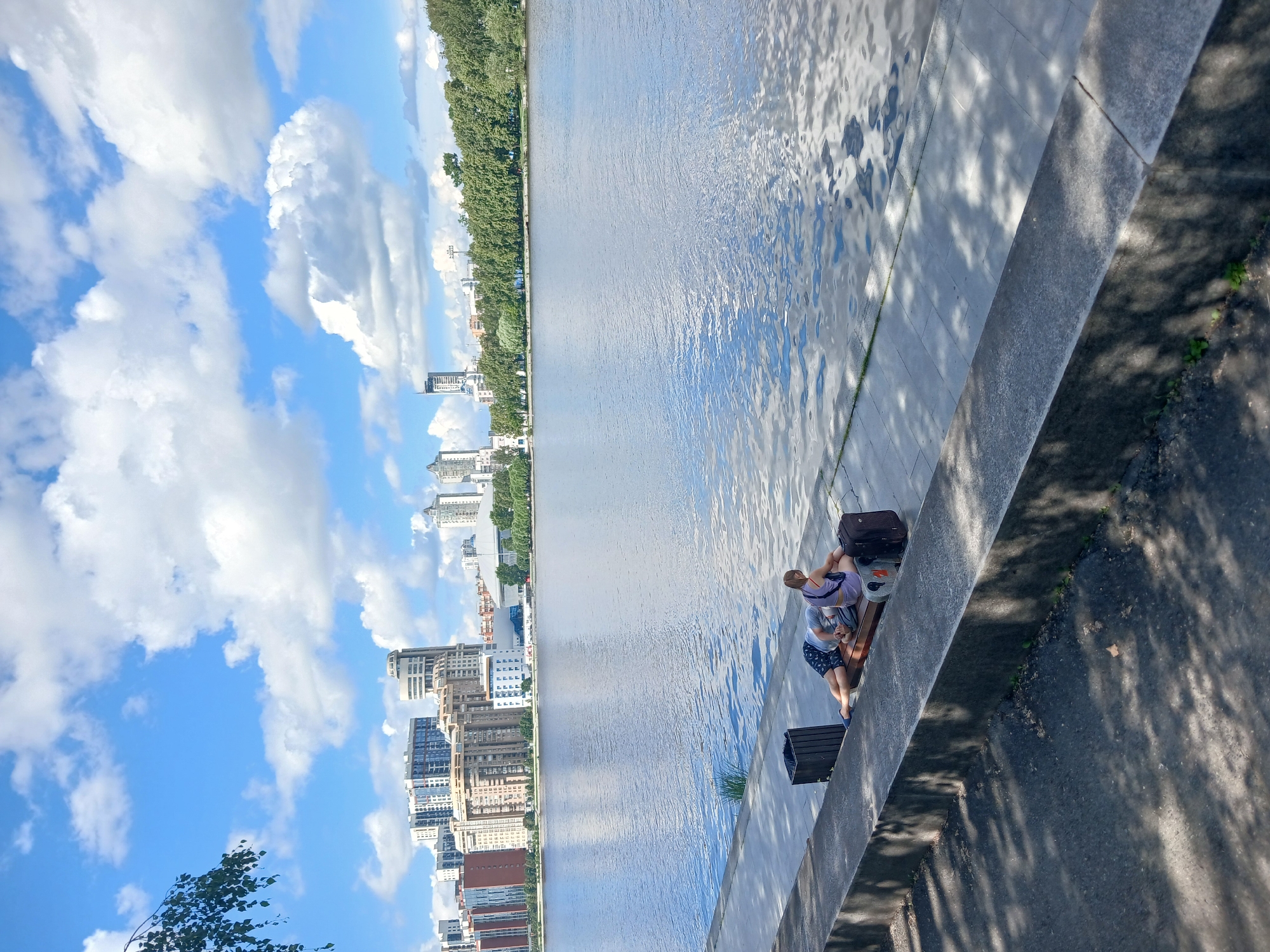 This image depicts a scene along the Iset River in Yekaterinburg, Russia, with the city skyline visible in the background. In the foreground, two individuals are resting on a park bench situated on a paved walkway adjacent to the river. A man, wearing dark shorts and a light-colored t-shirt, is lying on his stomach on the bench, his head propped on his arms. Beside him, a woman, dressed in a light purple top and shorts, sits on the bench, facing away from the viewer. Two suitcases, one dark and one lighter colored with orange accents, are placed on the ground next to the bench. A dark trash can is visible behind the man. The river, reflecting the blue sky and white clouds, occupies the middle ground. The background features a dense line of trees on the far bank of the river, beyond which are several modern buildings, including high-rises, characteristic of a city. The sky is bright blue with scattered cumulus clouds. The ground in the immediate foreground is asphalt, shaded by trees, with dappled sunlight filtering through.