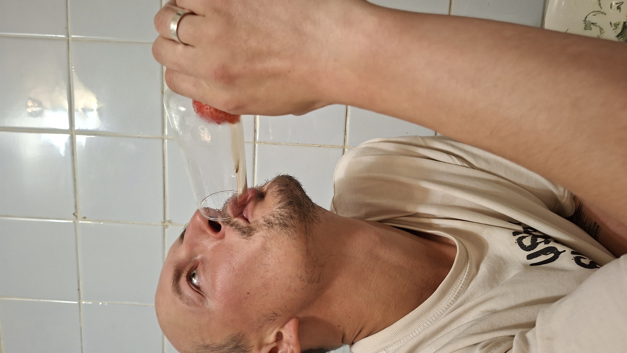 The image depicts an adult male drinking from a clear glass containing a red strawberry and a light-colored liquid. The scene is located in Забалоцкі сельскі Савет, Belarus. The man has a bald scalp, dark facial stubble, and is wearing a light-colored t-shirt with a dark graphic. A silver-colored ring is visible on his right ring finger, which holds the glass. His head is tilted back, with his gaze directed upwards, and his lips are pursed around the glass rim. The background consists of light-colored, possibly white, ceramic wall tiles with visible grout lines, and a portion of a white ceramic basin with a green pattern is visible in the lower right.