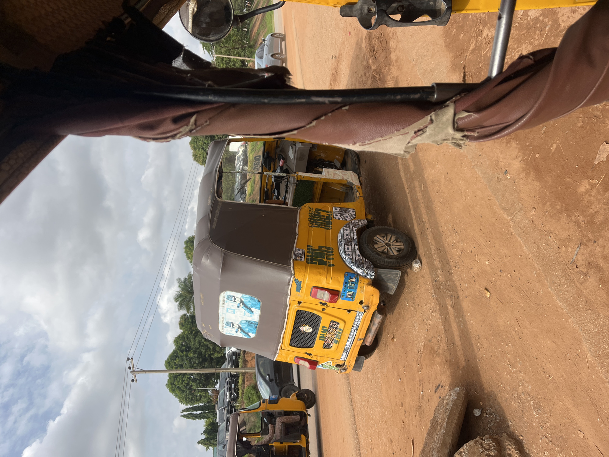 An outdoor, medium shot captures a yellow and brown auto-rickshaw parked on a dirt road in Galadimawa Village, Nigeria. The auto-rickshaw is decorated with images of men in blue shirts and pictures of money. In the background, other vehicles and trees are visible. The sky is partly cloudy. The image is taken from inside another auto-rickshaw, showing a torn brown seat in the foreground.