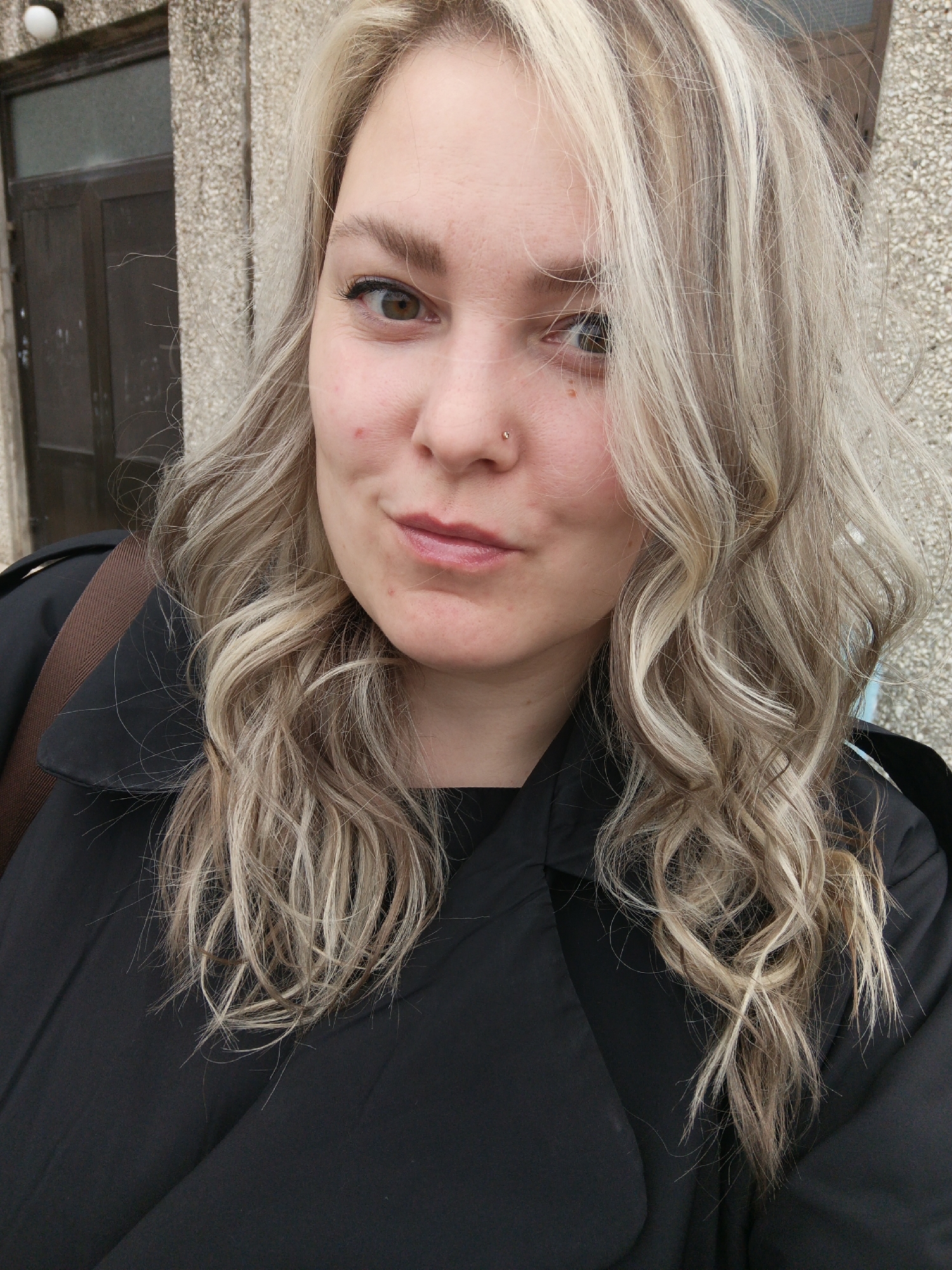 A woman with blonde, wavy hair is facing the camera. She has a nose piercing and is wearing a black coat. A brown bag strap is visible over her shoulder. Behind her is a building with a dark door and a textured wall. The location is Марковский, Russia.