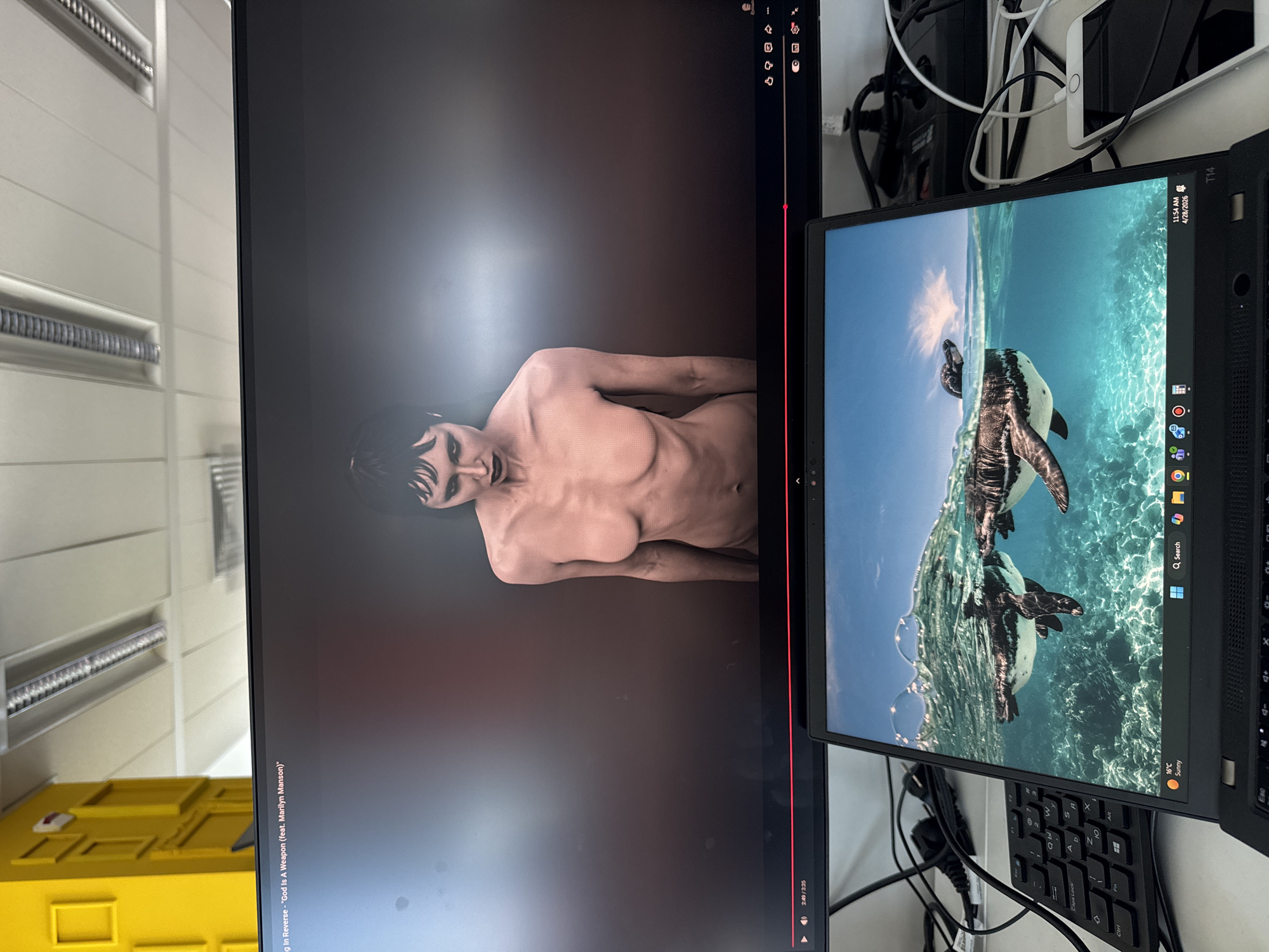 This media file captures a two-monitor desk setup in what appears to be an office or home environment in Sofia, Bulgaria. The image is taken at an angle, showing the screens upright but the camera's orientation is sideways.
The larger, primary monitor on the left displays a video, likely a music video, featuring a person with dark hair and a gaunt appearance, depicted from the waist up. The person is looking slightly to the left, and their visible torso shows pronounced ribs. The video appears to have a dark, dramatic aesthetic. Below the video, a progress bar indicates the current timestamp as 2:49 out of a total duration of 3:35. Visible text on the screen identifies the content as "g In Reverse - 'God Is A Weapon (feat. Marilyn Manson)'". Standard video player controls such as play/pause, volume, and settings icons are also visible along the bottom edge.
To the right, a smaller laptop, identified as a "T14" model, is open and displaying its desktop. The screen features a captivating wallpaper of a penguin partially submerged in clear blue water, swimming upwards. The laptop's taskbar is visible at the bottom, showing various application icons, a search bar, and a weather widget indicating "16°C Sunny". The system clock displays "11:54 AM" and the date "4/28/2026". The keyboard of the laptop is partially visible, with keys like "Caps" and "Esc" discernable.
The area around and between the screens is somewhat cluttered with various wires, cables, and charging devices, including a white tablet or smartphone mounted vertically. In the background, to the left, a light-colored wall or partition with horizontal grooves is visible, possibly part of an office cubicle or shelving unit. A bright yellow storage container is also partially visible at the bottom left of the frame.