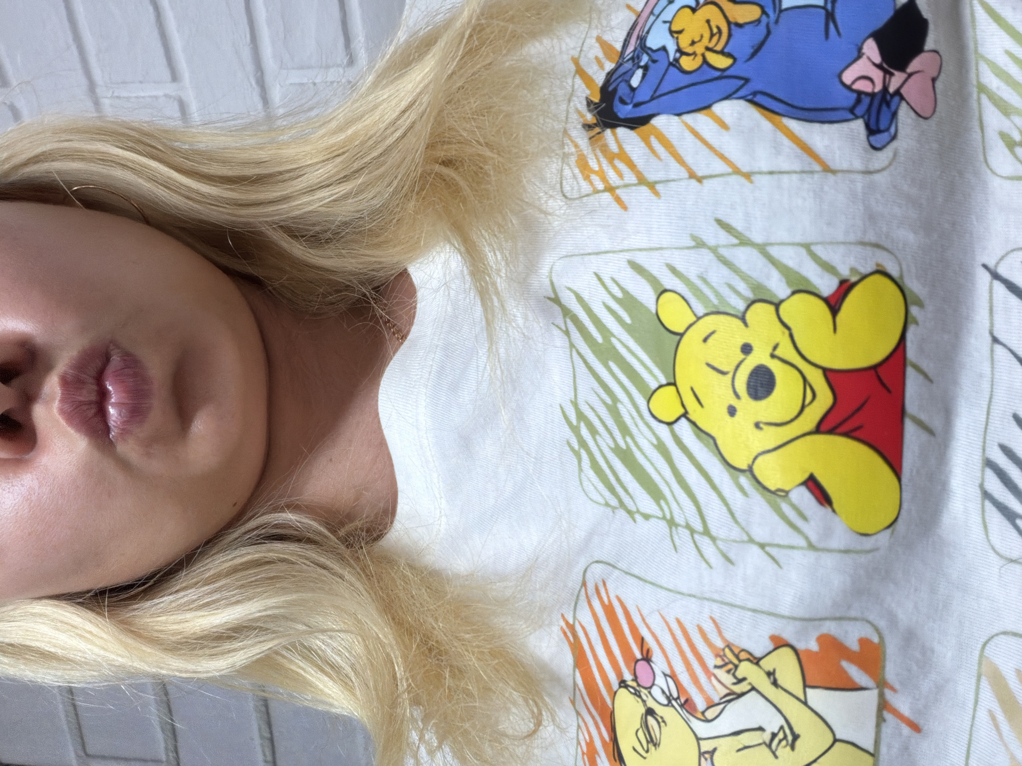 The image shows a close-up of a woman's face and upper torso, wearing a white t-shirt featuring cartoon characters. Her blonde hair is styled in loose waves, and her mouth is pursed, as if blowing a kiss. She is wearing a thin gold necklace. The t-shirt has three panels with illustrations: on the left, a yellow character with an orange striped background; in the center, Winnie the Pooh in a red shirt; and on the right, a blue donkey-like character with a pink bow. The background appears to be a white brick wall. The location of Chaikovsky, Russia is not evident in the image.