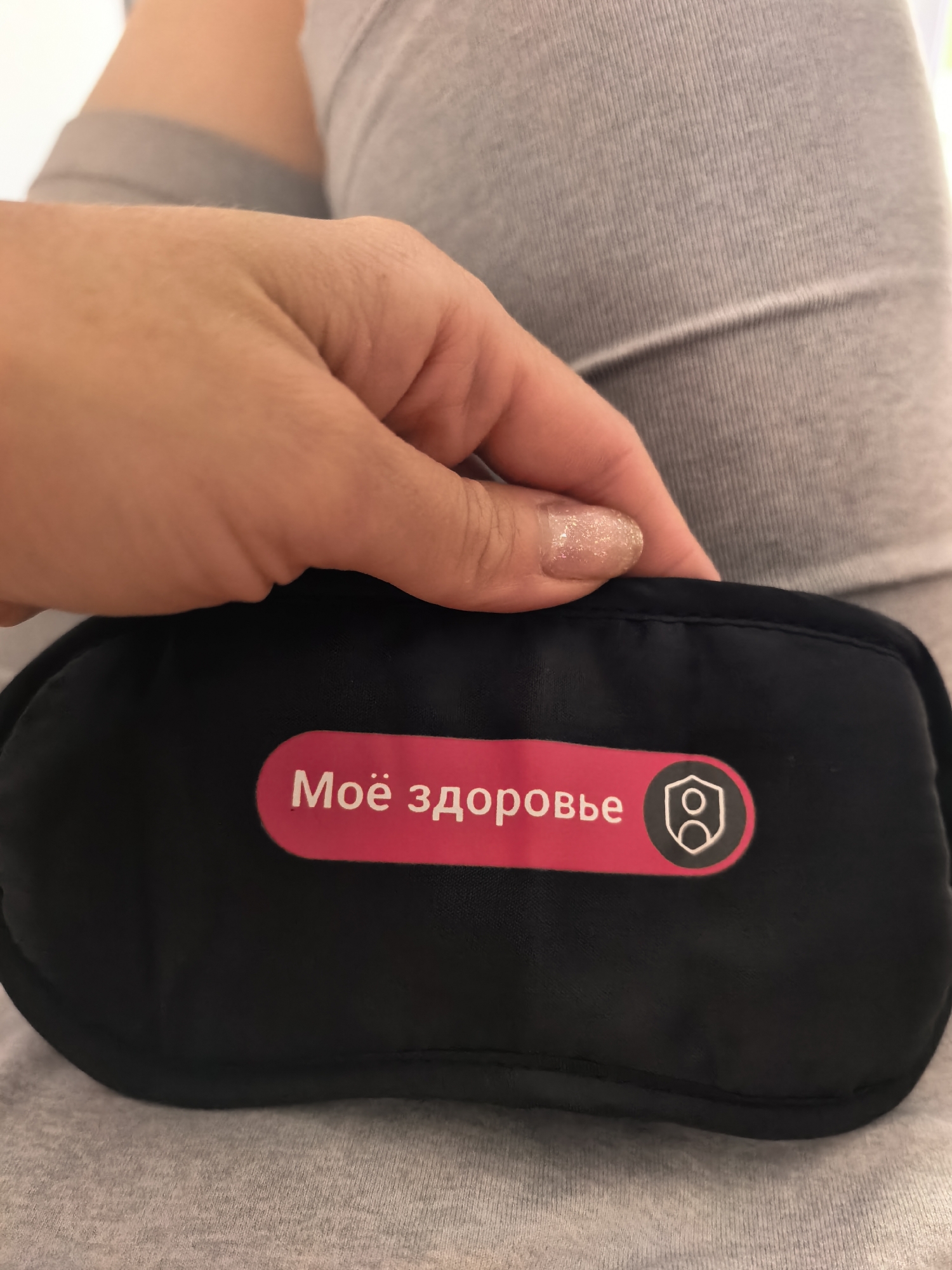A close-up shot shows a hand with glittery fingernails holding a black eye mask. The eye mask has a pink rectangular label with white Cyrillic text that reads "Моё здоровье" (My health) and a white shield-like icon with two figures inside. The background consists of a person's gray ribbed fabric clothing, possibly pants or a skirt. The image is framed such that the eye mask and the hand are the primary focus. The environment appears to be indoors, with soft lighting. No specific location in Sterlitamak, Russia can be determined from the image.