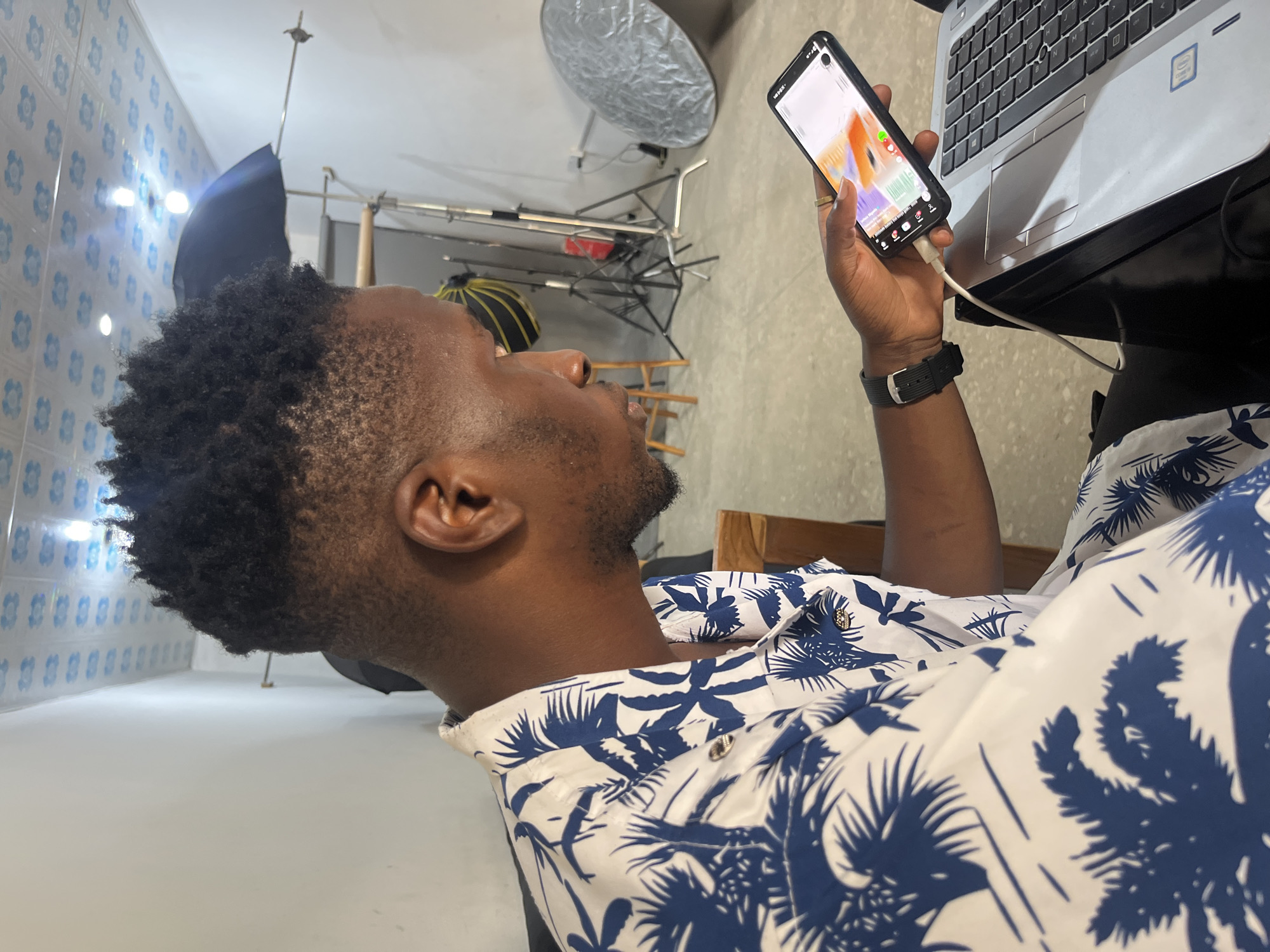 A Black male with short, textured dark hair and a goatee is seated, looking at a smartphone in his right hand. He wears a white short-sleeved shirt with a blue palm leaf pattern and a black wristwatch. The phone, connected to a silver laptop by a white cable, displays an image with orange and purple hues. The laptop is open, with its screen visible. The background suggests a studio or storage area, with lighting equipment including stands, a black umbrella, and a silver reflector. A wooden chair and a portion of another chair are visible. The ceiling is tiled with a blue and white pattern. The scene takes place in Gura Top, Nigeria.