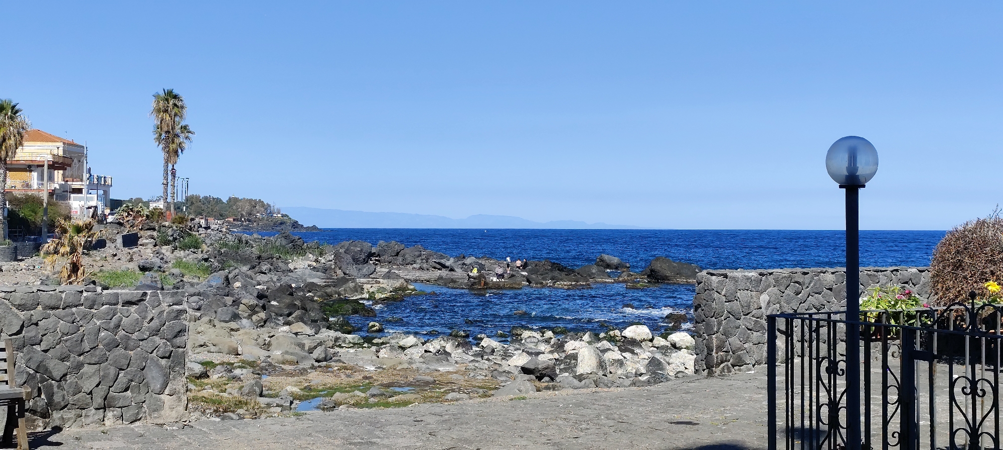 The image depicts a coastal scene in Aci Castello, Italy. A low, stone wall built from dark, irregular blocks runs across the lower left and center of the frame. Beyond the wall, there's a rocky coastline where dark volcanic rocks meet the blue water of the sea.
A group of people is visible on the rocks further out in the water. They appear to be casually positioned, perhaps exploring the coastline or relaxing. In the background, distant mountains are visible on the horizon under a clear blue sky.
On the right side of the image, another section of the stone wall frames the view. To the right of the wall, there is a black metal fence with decorative elements. Next to the fence, there's a street lamp with a spherical glass cover, and a bush with flowers. The foreground shows a paved area, likely a walkway.
To the left, buildings and palm trees add to the coastal cityscape of Aci Castello.