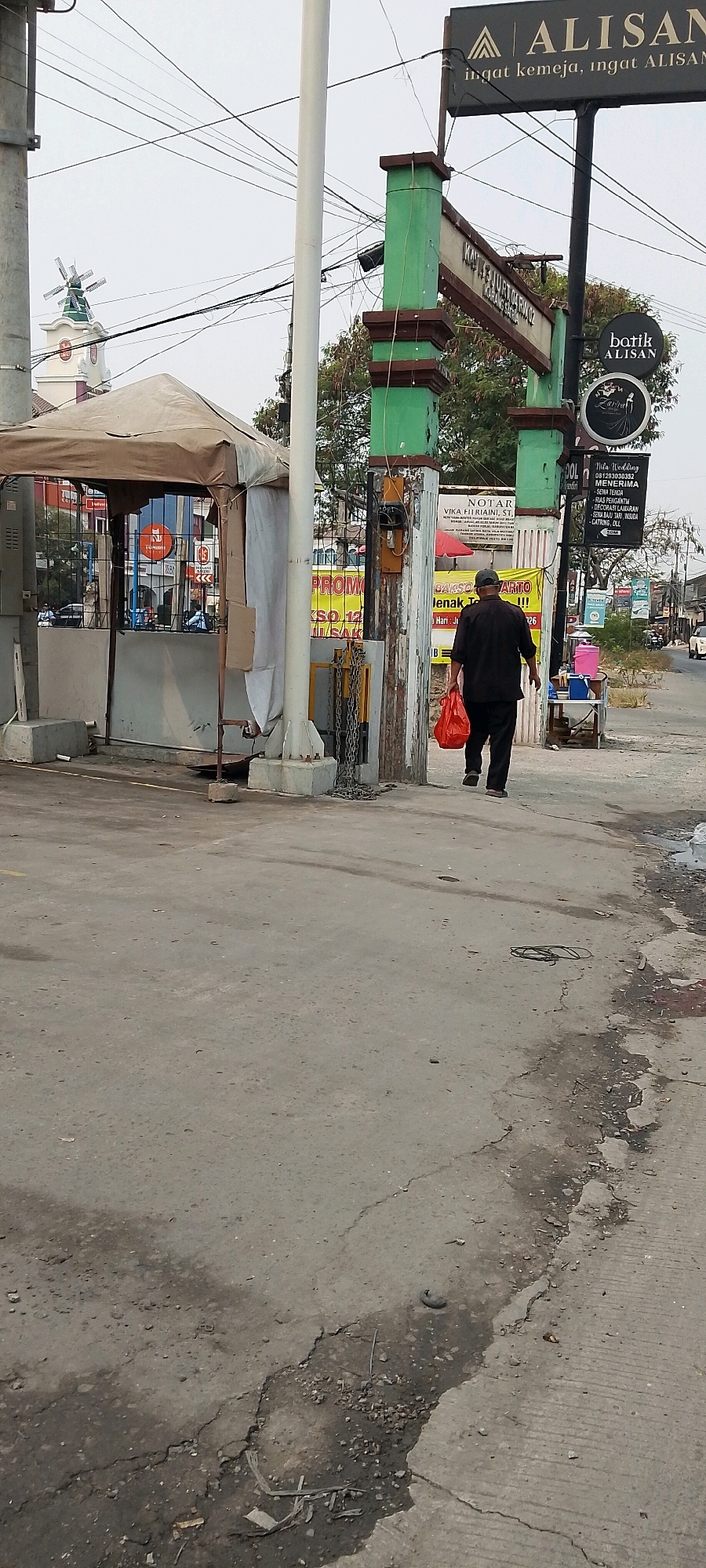 A man is walking away from the camera on a cracked concrete pathway. He is wearing a dark short-sleeved shirt and dark pants, and he is carrying a bright red plastic bag in his right hand. To his left, there is a guarded booth with a tan canopy and a white curtain partially covering the entrance. A chain hangs from a post to the right of the booth. Further to the right, an archway structure with green posts and dark trim stands, bearing signs. One sign, a black circle with white text, reads "batik ALISAN". Another sign above it is partially obscured but appears to be a dark rectangle with white text. A tall white pole stands to the left of the archway. In the background, trees, buildings, and overhead wires are visible. To the far left, a white tower with a Dutch-style windmill on top is present. The scene appears to be on a street in Karangsatria, Indonesia.