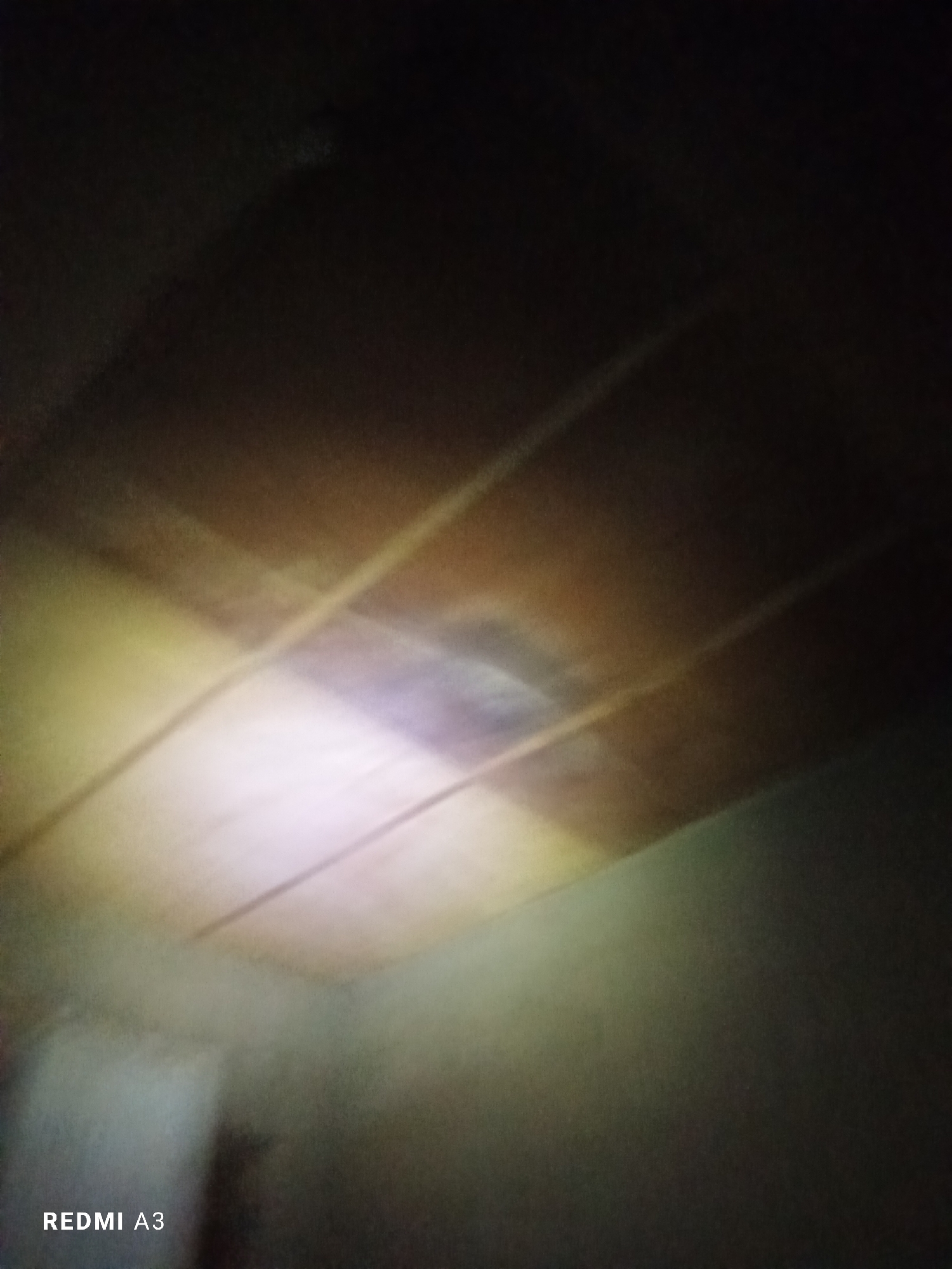 This is a dark, out-of-focus image that appears to be taken indoors. The primary subject is the ceiling, which is made of wooden planks and shows some discoloration or a stain in the center, illuminated by a bright light source creating a rainbow-like effect. There are also diagonal lines of light across the ceiling, suggesting a patterned illumination or reflections from something outside the frame. The walls are visible at the bottom of the image and are a muted, hazy green or gray color. The overall atmosphere is dim and somewhat mysterious due to the low light and blurriness. There is no discernible activity or event taking place. The visible text in the lower left corner reads "REDMI A3", which is likely a camera or phone model identifier. The time of day and weather are not discernible.