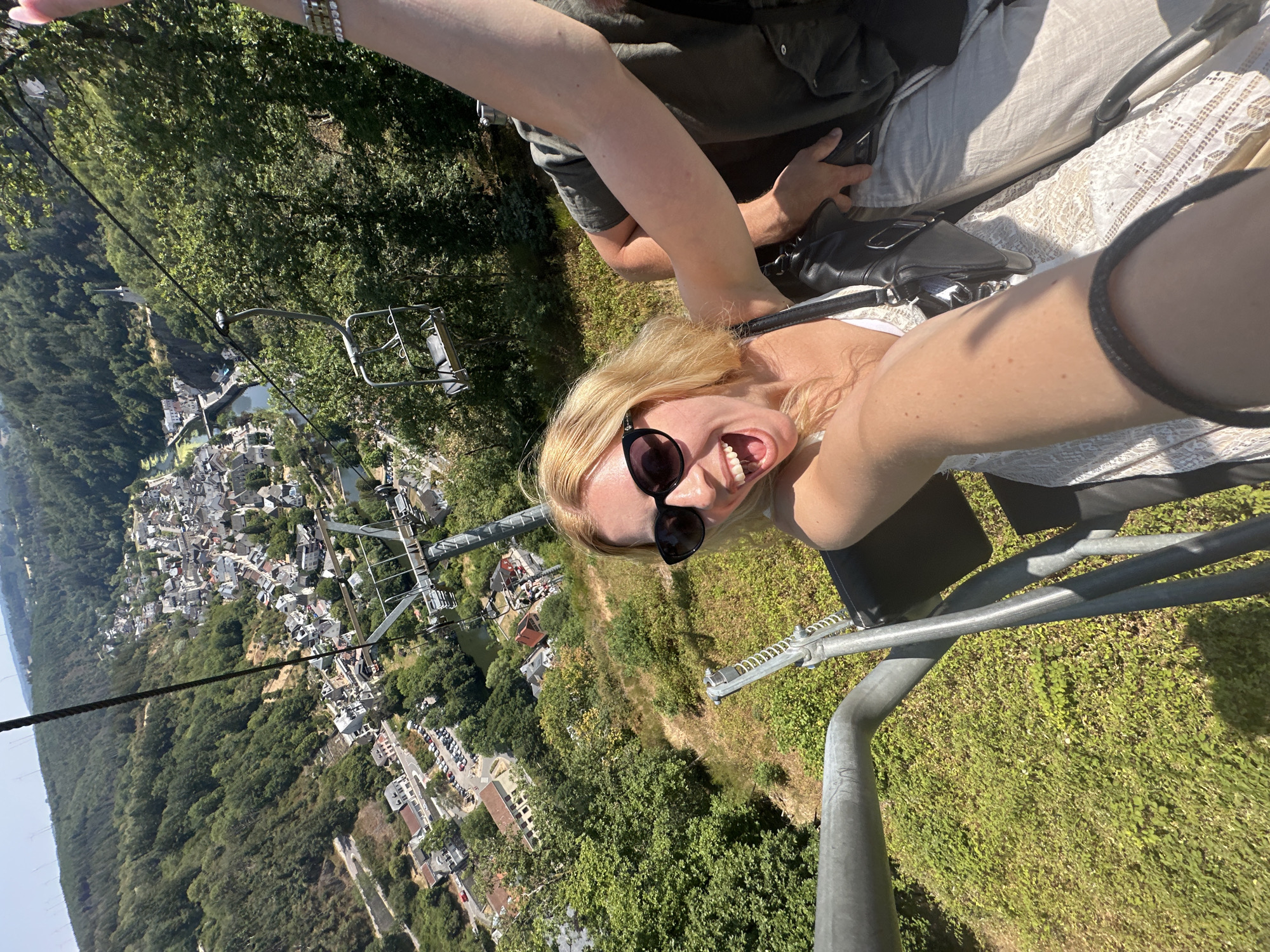 The image shows a woman in a chairlift, smiling broadly and looking at the camera. She has blonde hair and is wearing sunglasses and a white lace dress. In the background, a town with red-roofed buildings is visible, nestled in a valley surrounded by lush green hills. A river runs through the town. The chairlift is moving over the town, offering a panoramic view. The scene appears to be in Vianden, Luxembourg, known for its medieval castle and picturesque landscape.