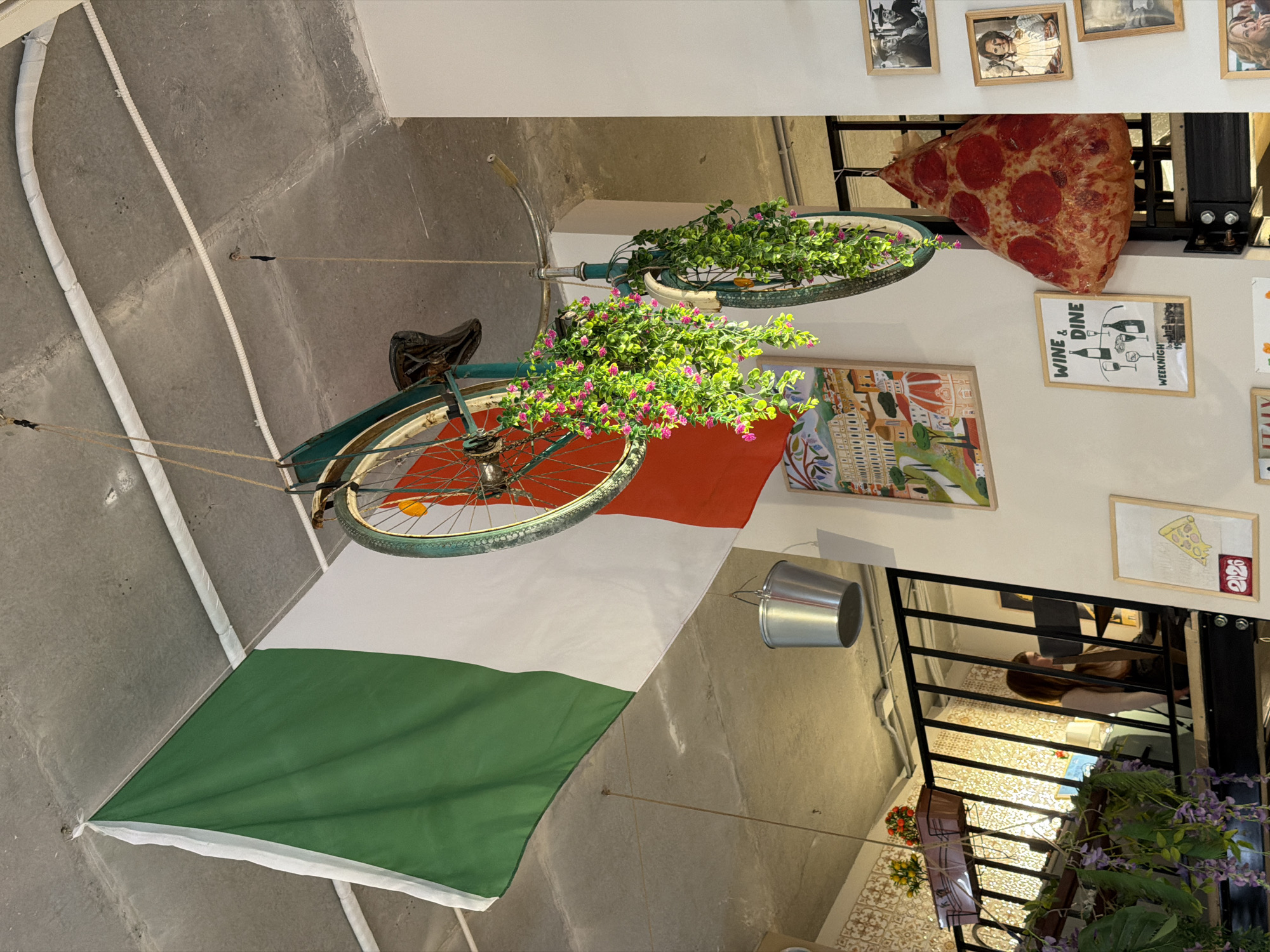The image depicts an Italian-themed restaurant or establishment. An old bicycle is suspended from the ceiling, decorated with artificial flowers in both wheels and draped with an Italian flag. Adjacent to the bicycle are framed pictures, including a pizza slice and a "Wine & Dine" advertisement. A decorative pizza slice is placed on the wall. A metal bucket is also hanging from the wall. A railing is visible, and a woman with long brown hair is sitting at a table on the other side of the railing. Further down, hanging plants decorate the area. Given the presence of the Italian flag and decorations, the location could be an Italian restaurant. However, there is no information in the image to suggest that the location is in the city of Izhevsk, Russia.