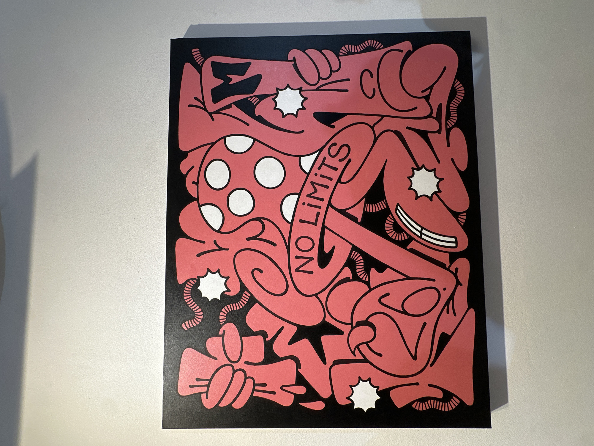 A framed artwork hangs on a light-colored wall. The artwork is rectangular and features a black background with a bold, graphic design in bright pink and white. The pink elements are abstract and organic, forming shapes that resemble figures or creatures in a chaotic, interconnected composition. Some elements are outlined in black. White circles are present on a large, dome-like pink shape, and white starburst shapes are scattered throughout the design. A banner with the words "NO LIMITS" in black lettering curves across the center of the artwork. The overall style is reminiscent of street art or graffiti. The wall appears to be textured. The lighting suggests an indoor setting, possibly a gallery or a commercial space. The artwork is positioned at eye level.