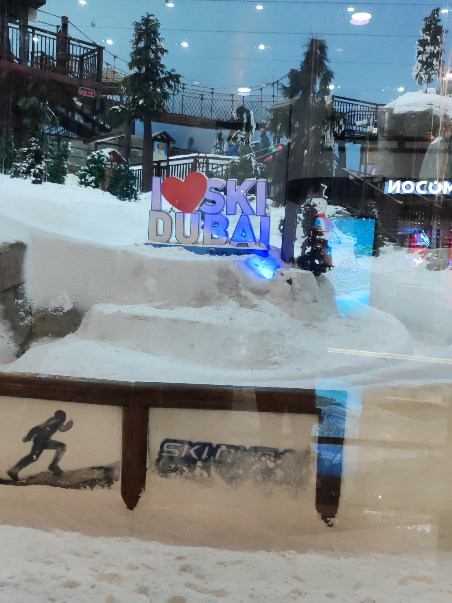 The image depicts the indoor ski slope attraction at Ski Dubai, located within the Mall of the Emirates in Dubai, United Arab Emirates. A large, three-dimensional sign displaying "I LOVE SKI DUBAI" in a blue, illuminated font is positioned in the foreground, with a red heart symbol in place of the word "LOVE." Surrounding the sign is a snow-covered artificial landscape with sculpted snow mounds and artificial pine trees. In the background, an elevated walkway with railings and stairs is visible, along with other elements of the indoor ski resort, such as ski lifts and netting. A digital clock displaying "20:37" is visible on a structure in the background. The scene is viewed through a glass barrier, which reflects some of the surrounding environment, including lights and what appears to be another indoor space. A portion of a wooden railing with two graphic panels is visible at the bottom of the image; one panel shows a silhouette of a skier or runner, and the other displays the word "SKI." The overall impression is an artificial winter environment within a commercial setting.