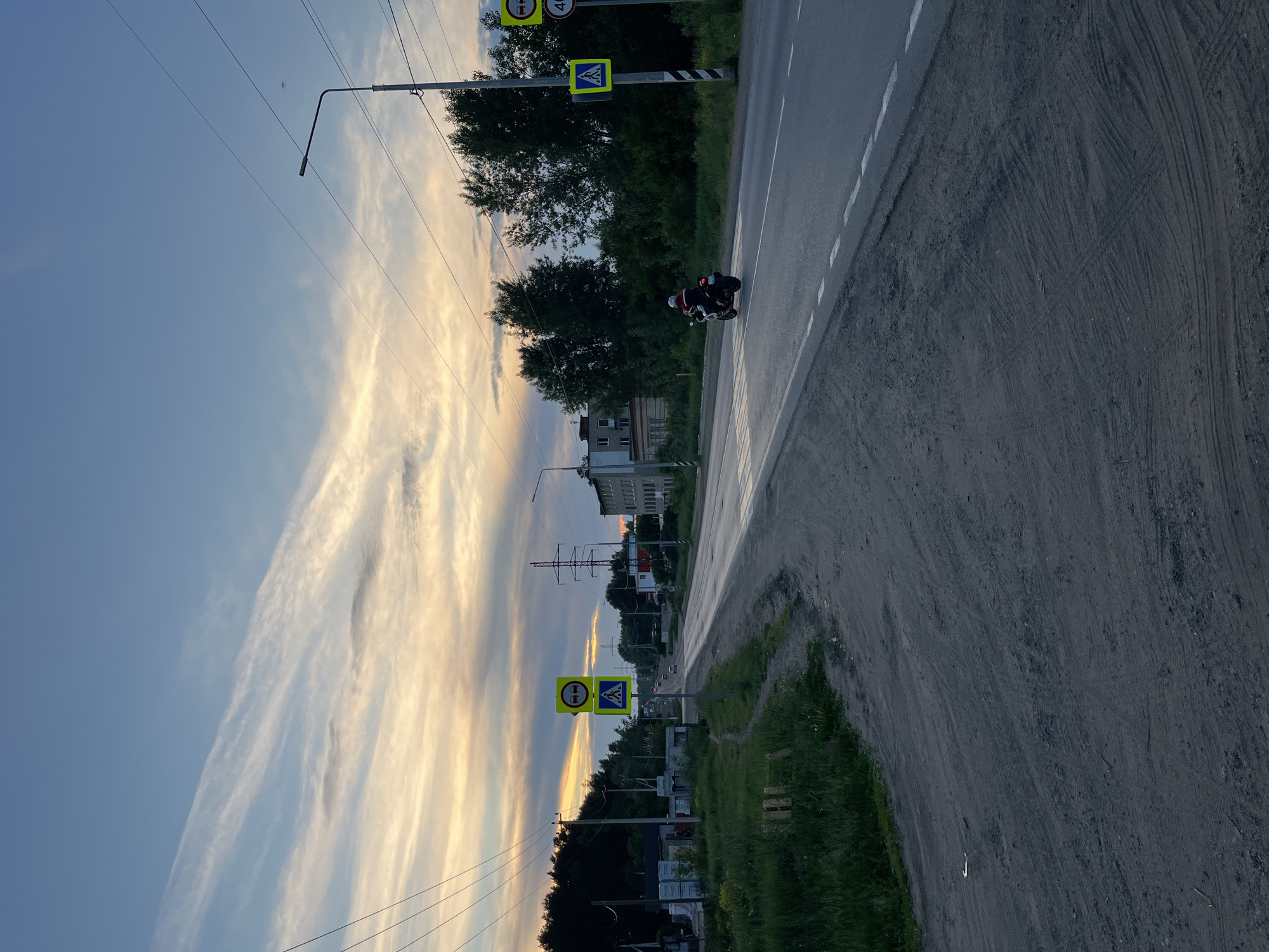 The image depicts a rural road in Chaikovsky, Russia, at dusk. The sky is a blend of blue, orange, and white, with wispy clouds suggesting the setting sun. A motorcycle is traveling on the right side of the asphalt road, which has white dashed lines marking its lanes. On the right side of the road, a lamppost stands tall, accompanied by traffic signs indicating a pedestrian crossing and a speed limit of 40 km/h. Trees line the road, with a few buildings visible in the distance, including a multi-story structure. The left side of the road features a dirt path, overgrown with grass and some construction materials. The overall scene is one of a quiet evening on a road leading out of or into a semi-urban area.