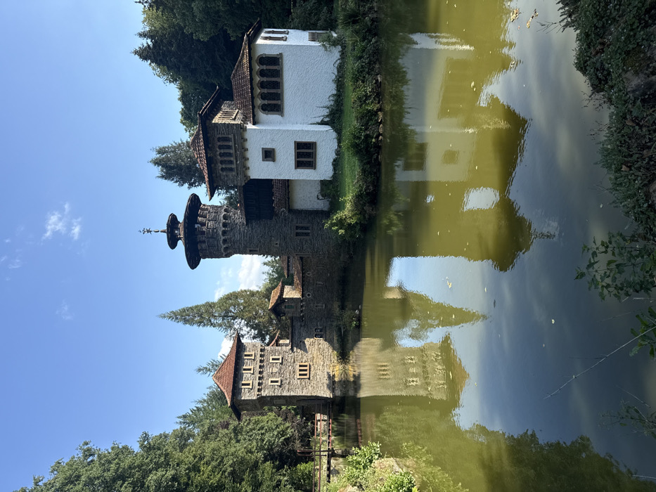 The image displays a multi-component, castle-like structure situated on the edge of a calm, greenish-brown body of water, likely a moat or pond, under a clear blue sky in Groussbus-Wal, Luxembourg. The building complex features a prominent, centrally located, cylindrical stone tower with a distinctive multi-tiered, circular roof structure and finial. To its right, a two-story, white-washed building with brown-tiled roofs and a mix of arched and rectangular windows connects. To the left of the central tower stands a square, stone-built tower with multiple small rectangular windows and a red-tiled gabled roof. This left tower connects to the bank via a small wooden bridge. The water surface clearly reflects the architectural elements. The structures are surrounded by dense green trees and foliage, forming a natural backdrop. No individuals or specific actions are visible within the scene.