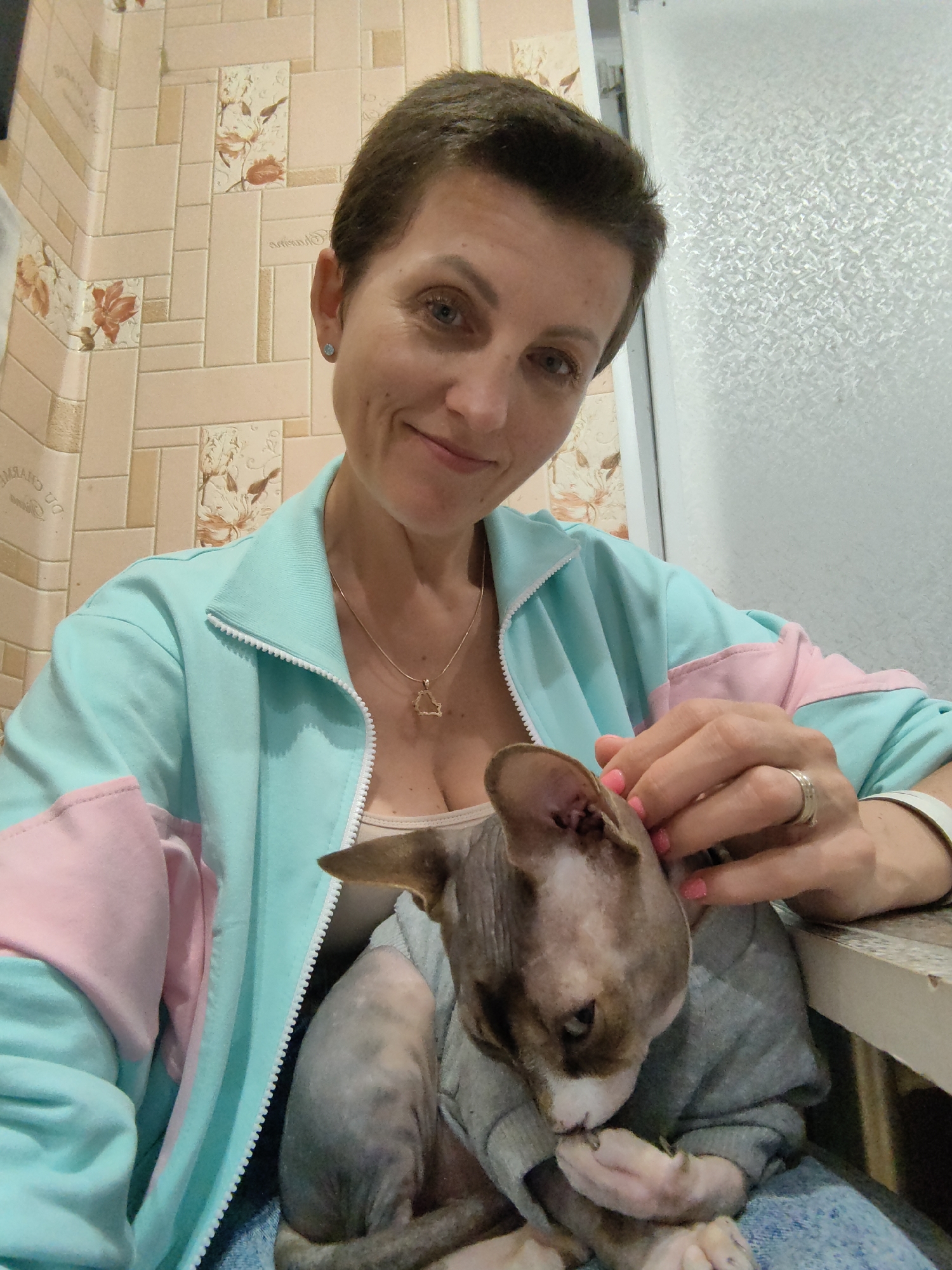 A woman with short brown hair and blue eyes is holding a Sphynx cat. She is wearing a light blue and pink tracksuit jacket and a necklace. The cat is wearing a grey sweater. The background appears to be a bathroom with tiled walls. The lighting is artificial. The image is a selfie taken by the woman. The location of Myadzyel, Belarus is not visually identifiable within the image.