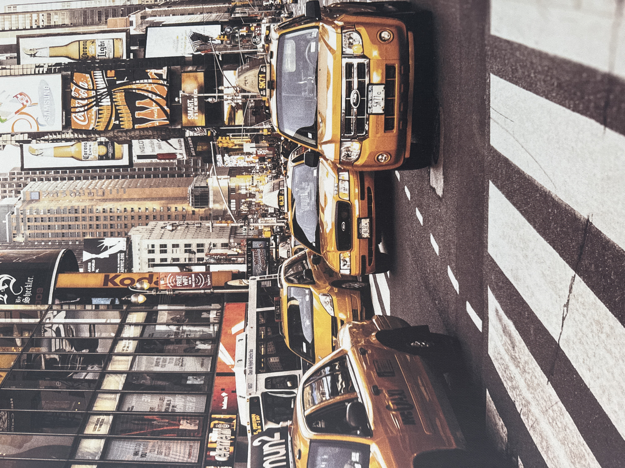 The image depicts a busy street scene in a city, characterized by multiple yellow taxis and tall buildings adorned with large advertisements. The foreground features a crosswalk with a yellow taxi prominently in the right foreground, displaying the license plate "5K71C". Another yellow taxi is visible behind it. To the left, another yellow taxi partially enters the frame. In the background, a white bus with the text "Clean Air Hybrid Electric Bus" is seen, along with more yellow taxis and a dense urban environment filled with illuminated billboards. The advertisements visible include brands like Coca-Cola, Corona Extra, and Samsung. Traffic lights are also present, indicating an active intersection. The overall impression is one of a bustling metropolis, likely Times Square in New York City, given the iconic yellow taxis and dense advertising. The provided location of Anudinovka, Russia, is not supported by the visual evidence in the image.