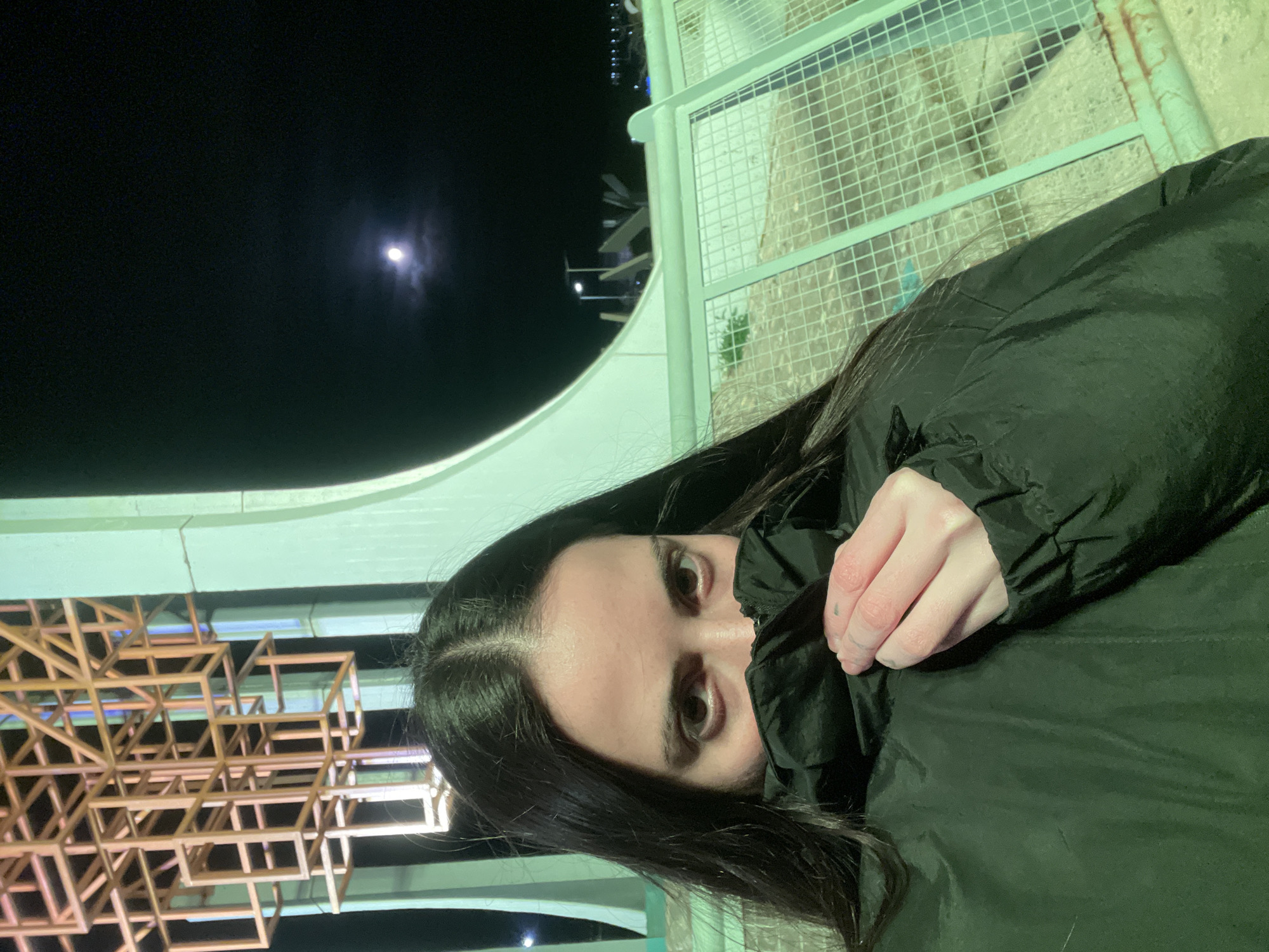 A person is shown at night, partially obscured by a black jacket held up to their face. The person has dark hair and light skin. Behind the person is a night scene with the moon visible in the upper left corner of the frame. The person appears to be near an architectural structure of wood or metal, constructed of intersecting lines. The background also shows portions of a structure with a fence-like design. The location is believed to be in the city of Ushuaia, Argentina.
