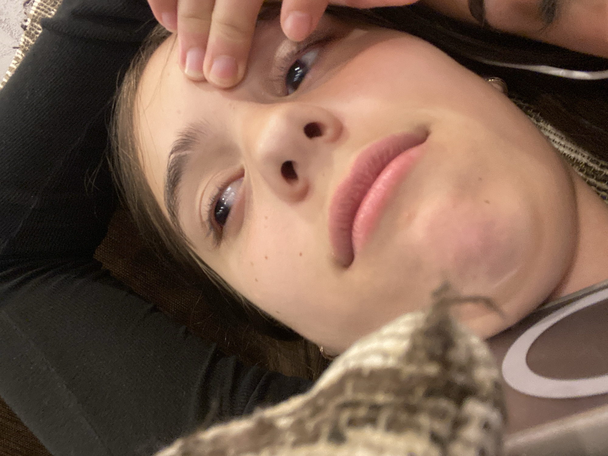 A person is lying down, taking a selfie with their head tilted. Their left hand is near their forehead, with fingers slightly touching the skin. The person has light skin, dark eyebrows, and dark eyes looking upwards. Lips are pink and slightly parted. A blurry object in a brown and white pattern is in the foreground. A dark-colored garment, possibly a sweater, is visible on the left. An earbud cord is visible near the right ear, indicating a possible headset. The location and surrounding environment are not identifiable from the image. The location in Dragovishtitsa, Bulgaria, cannot be determined based solely on the image.