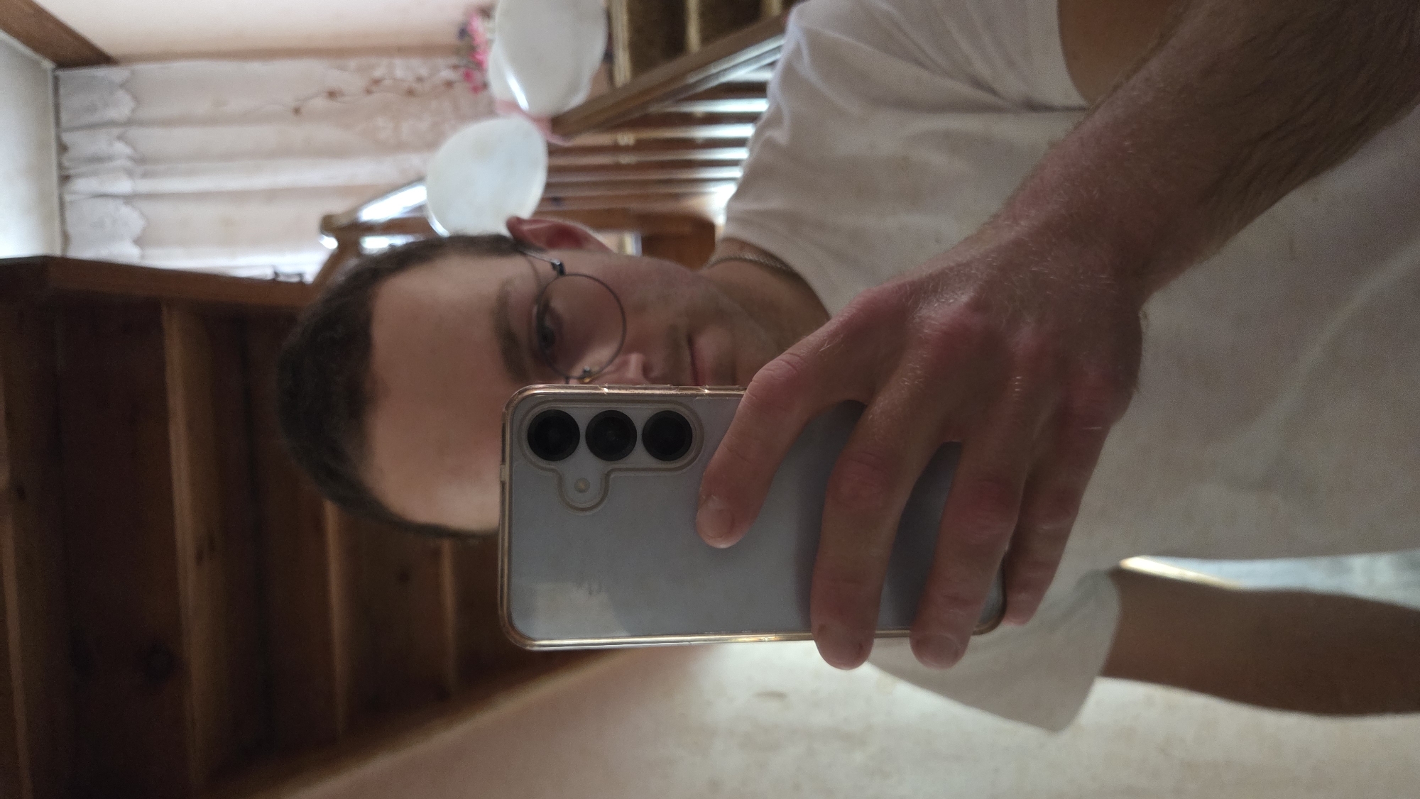 A man wearing glasses is taking a selfie in a mirror. He is wearing a white t-shirt and holding a light blue smartphone with three camera lenses. The reflection in the mirror reveals a wooden staircase and white balloons. The location is Unknown, Unknown.